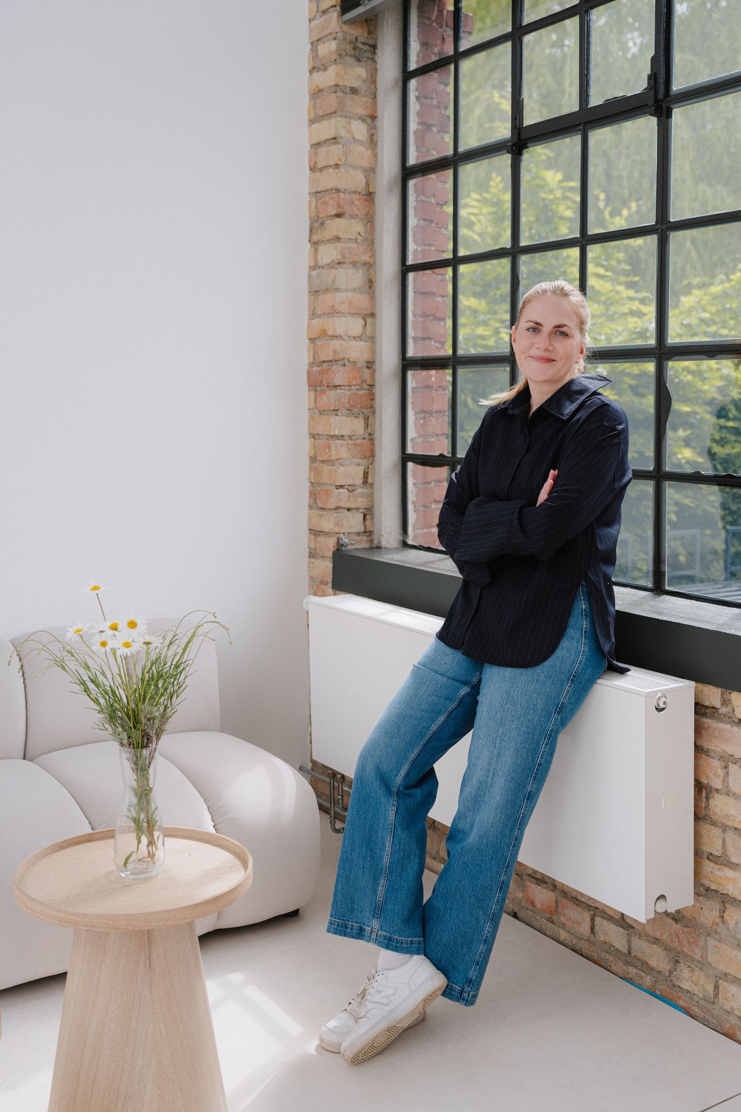 Woman in black shirt and jeans leaning against window sill in modern loft space with brick walls and white sofa.