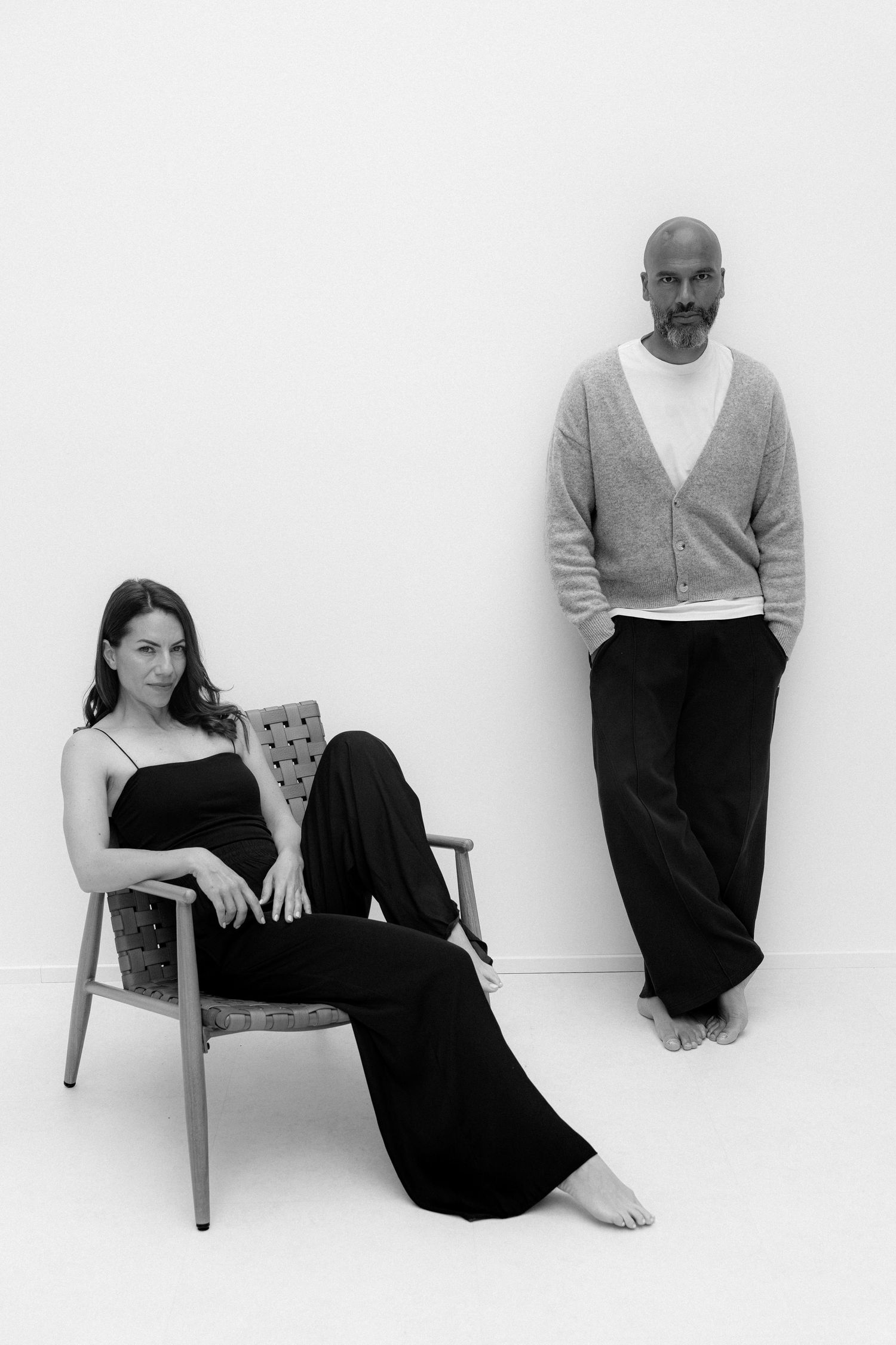 Black and white photo of a woman in black sitting in a chair and a bald man in a cardigan leaning against a white wall.