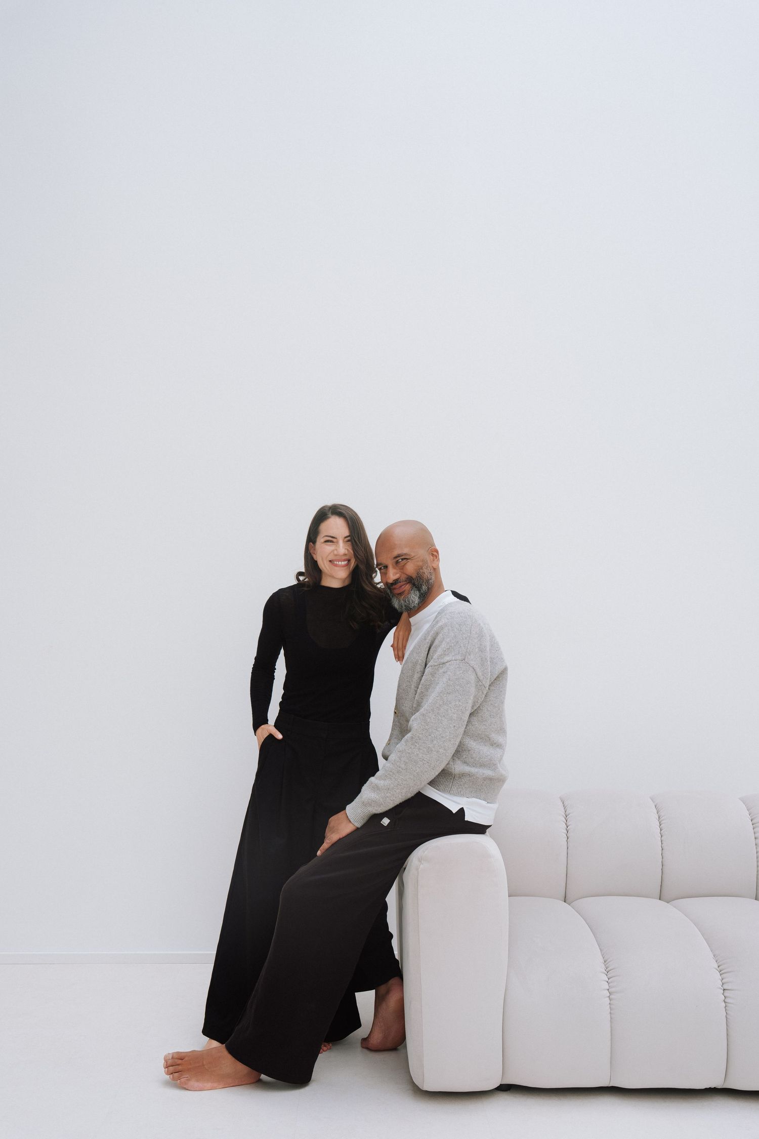 A woman in black dress and a man in gray cardigan pose together beside a white sofa against a white background.