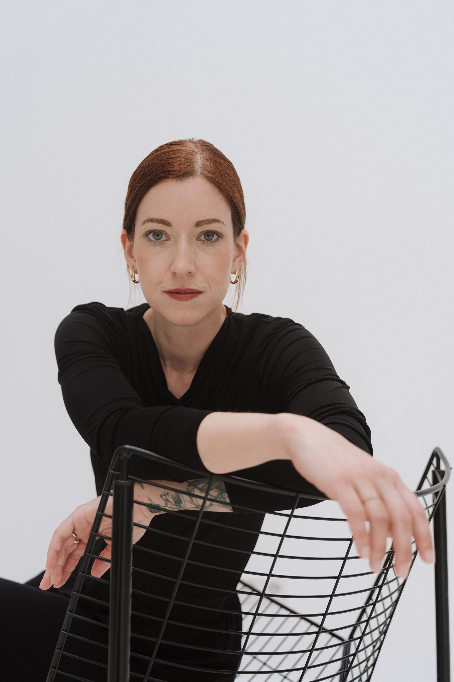 Woman in black top with tattoos leans on wire chair, posing confidently against white background.