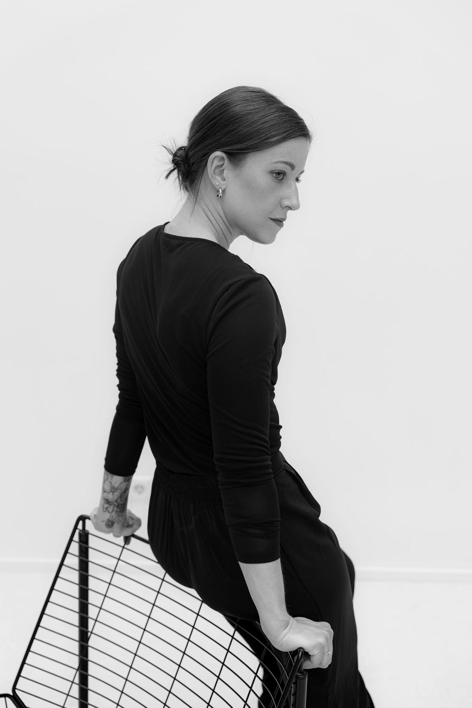 Black and white portrait of a woman in a black dress sitting on a wire chair, looking over her shoulder.