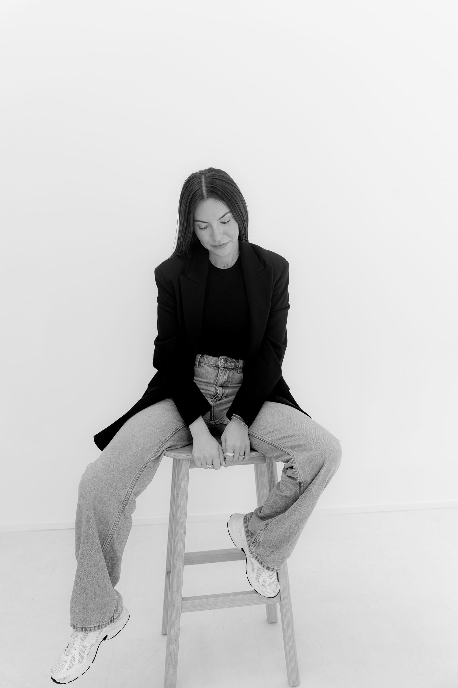 Black and white photo of a woman in blazer and jeans sitting on a stool against a white background.