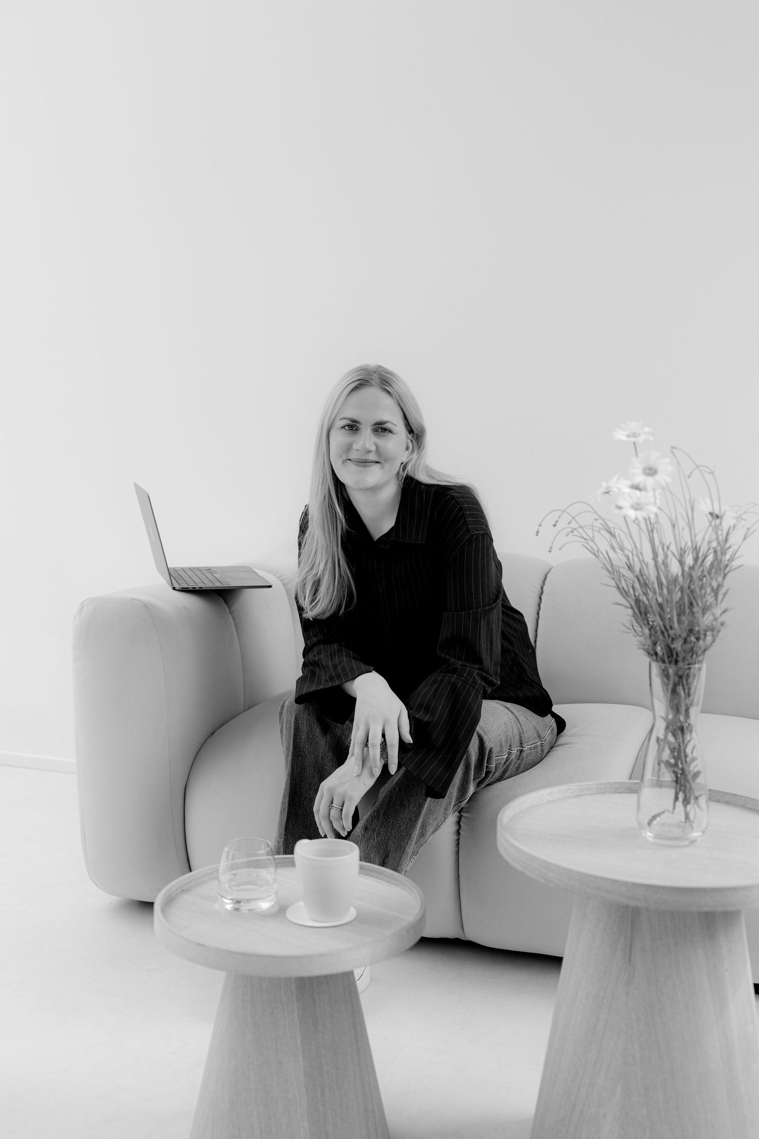 Black and white photo of a smiling blonde woman sitting on a modern sofa with a laptop, small tables, and a floral vase nearby.