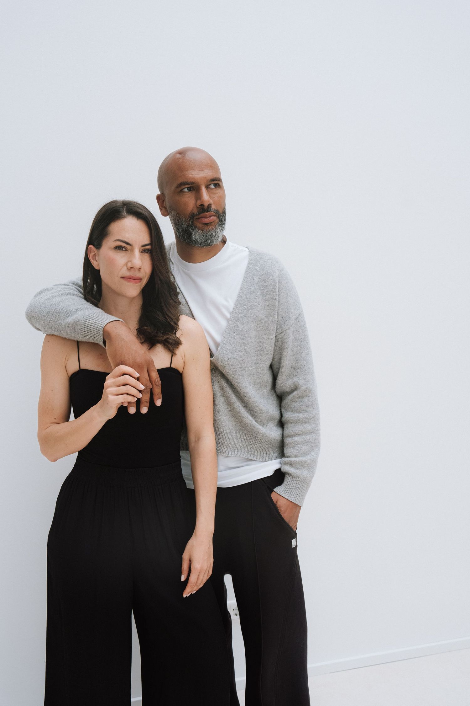 A man and woman in black and gray outfits pose together against a white wall, the man resting his arm on her shoulder.