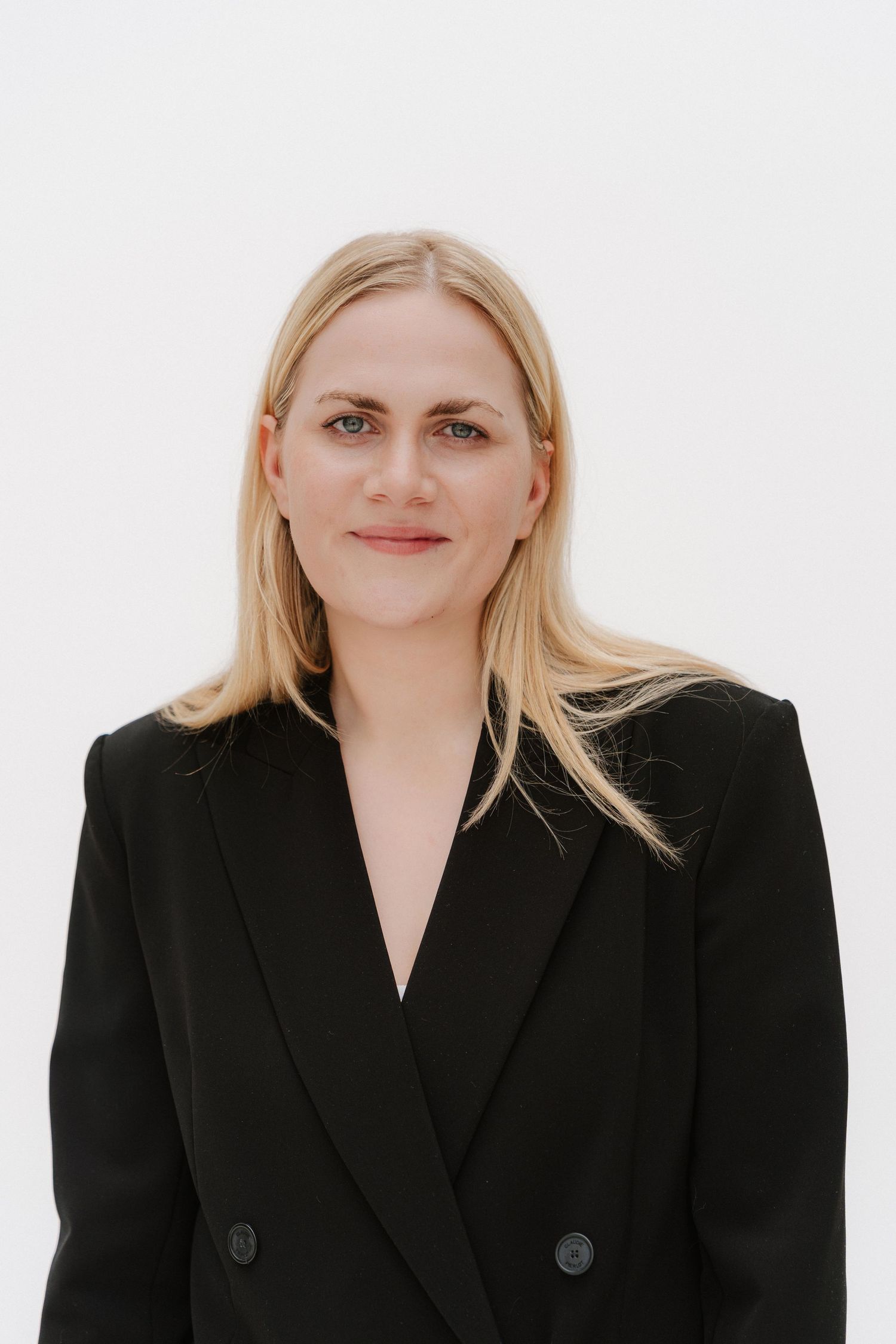 Professional blonde woman in black blazer posing against white background for corporate headshot.