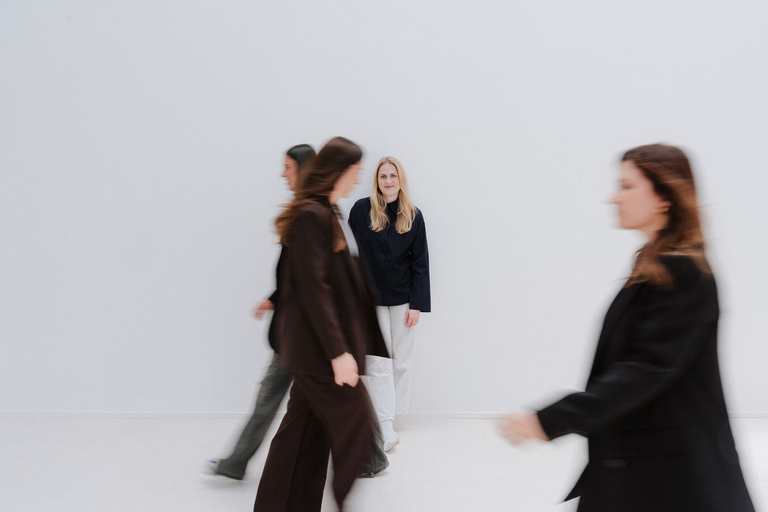 A woman stands still against a white wall while blurred figures in dark clothing walk past her.