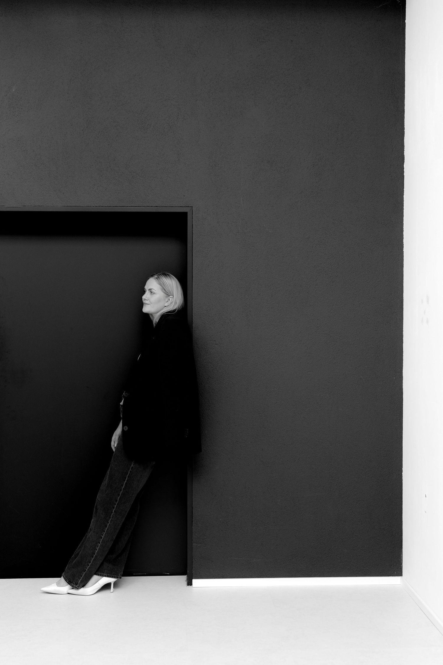 Black and white photo of a blonde woman in dark outfit leaning against a black wall panel on white floor.
