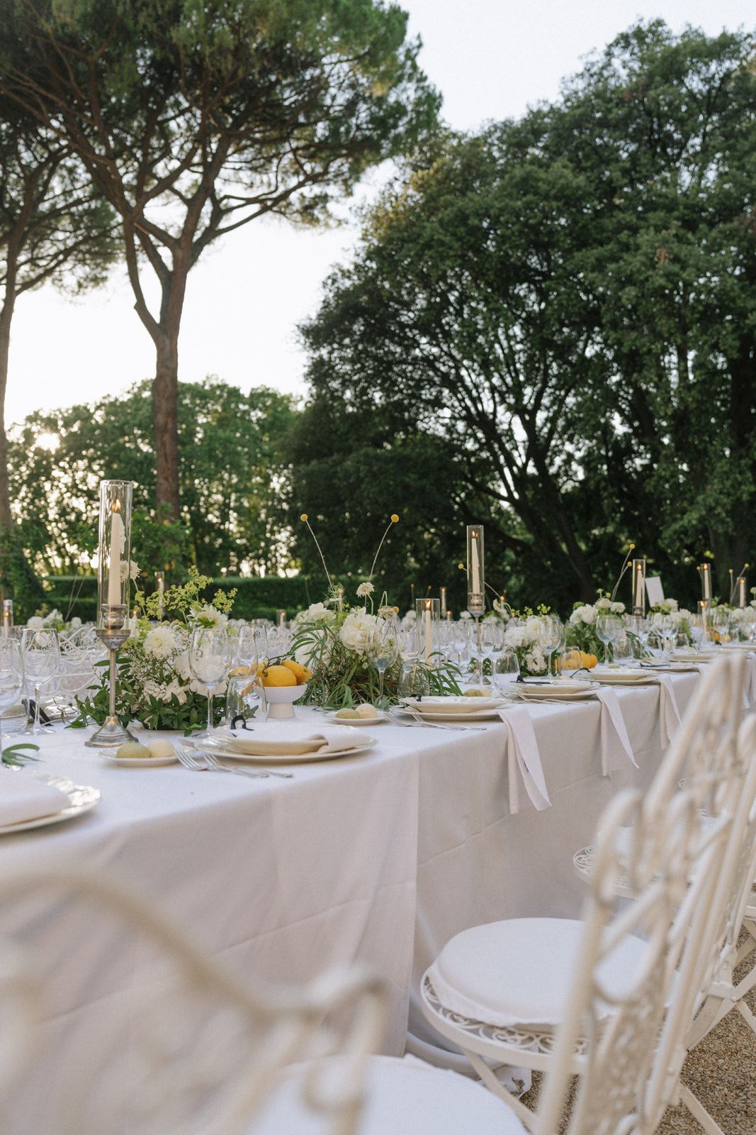 Elegant outdoor wedding reception table set with white linens, floral centerpieces, candles, and ornate chairs in a lush garden.