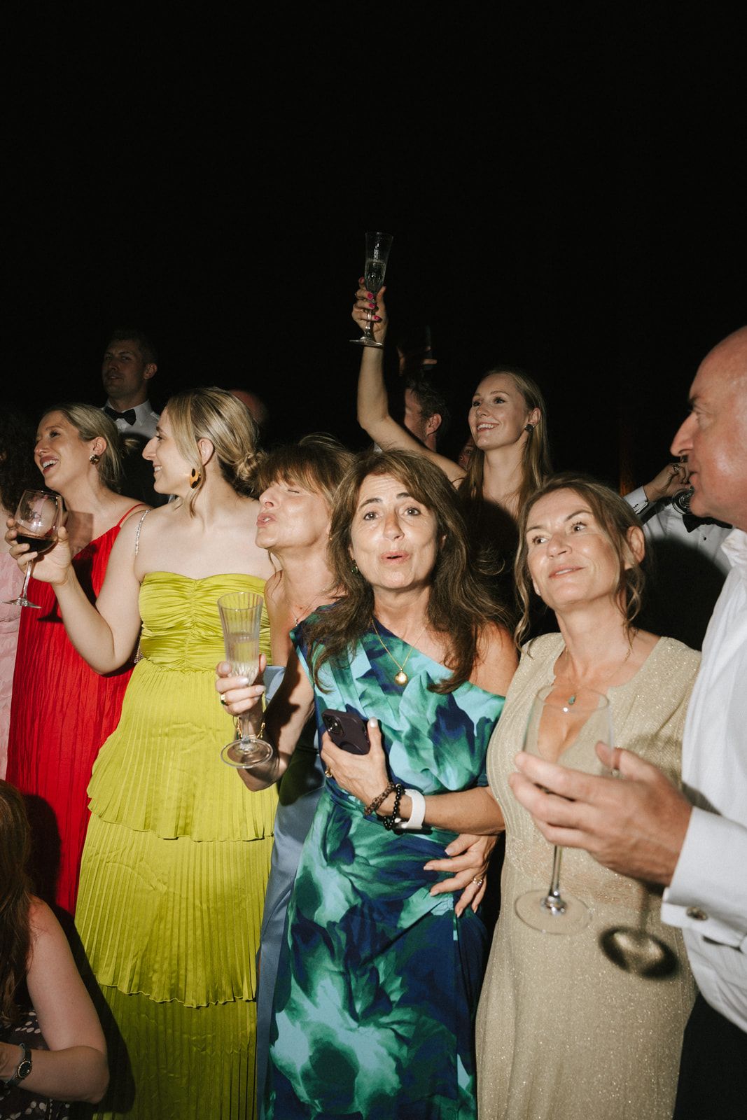 Guests celebrating at a party, holding champagne glasses and smiling, dressed in colorful formal attire.