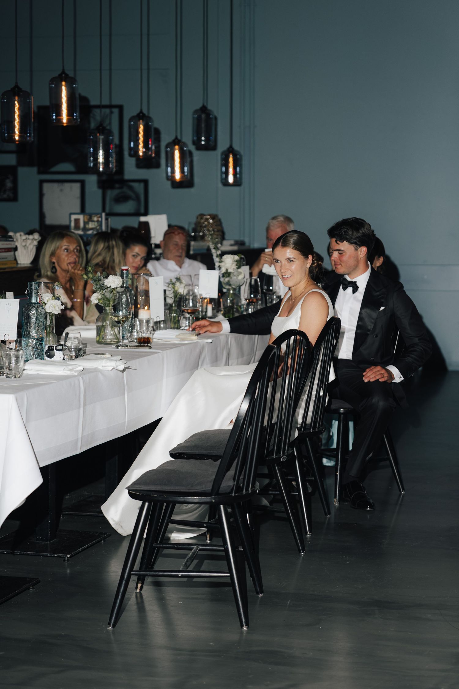 Modern reception venue with pendant lights and guests seated at black chairs with white tablecloths.