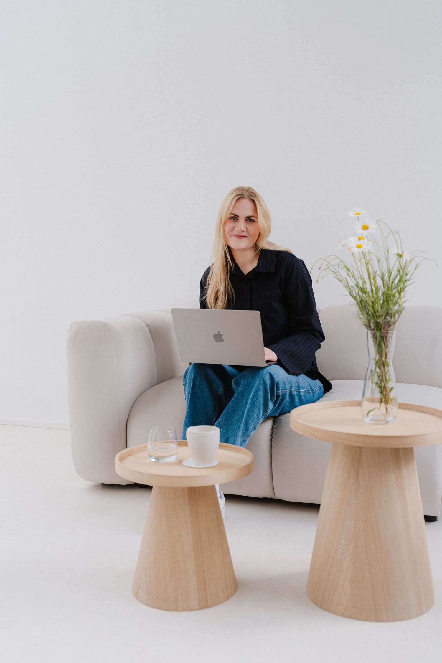 Blonde woman sitting on a modern sofa with a MacBook laptop, beside wooden side tables with flowers and a coffee cup.