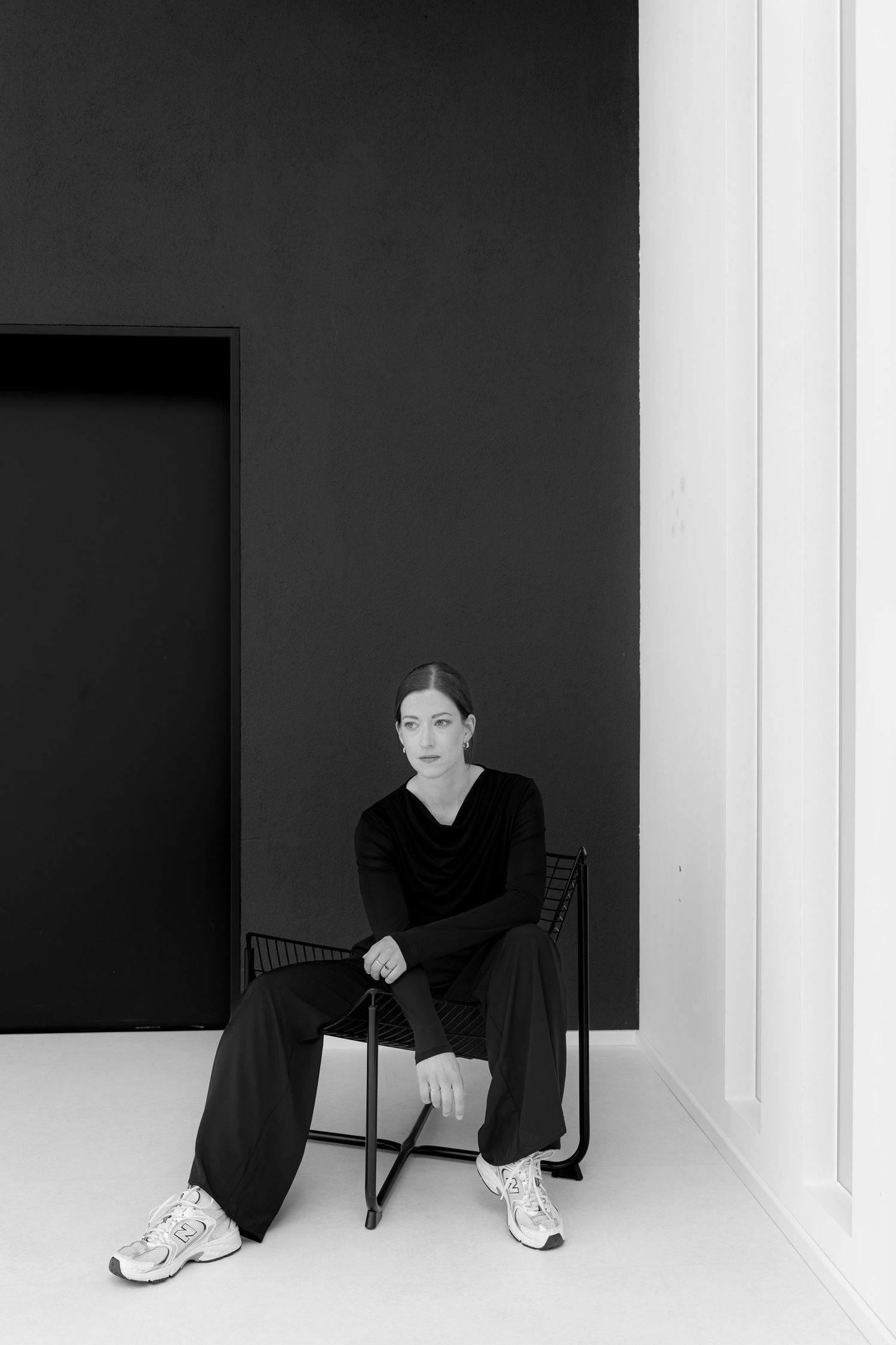 Woman in black outfit sitting on a minimalist chair against a dark wall in a black and white photo.