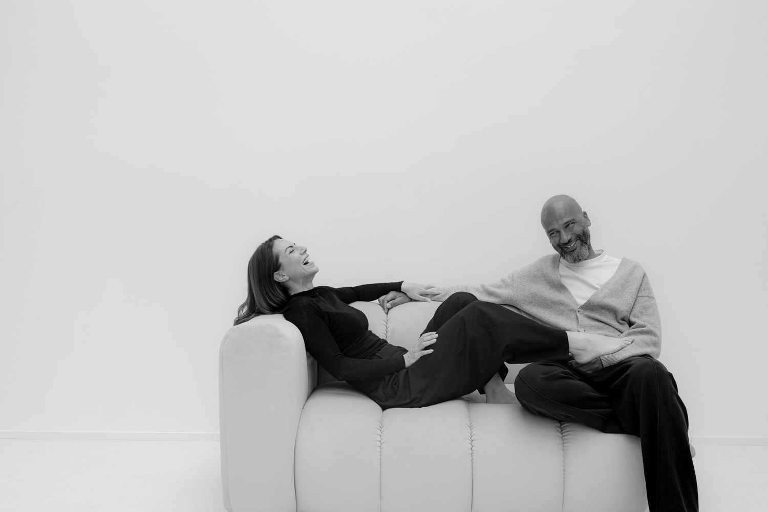 Black and white photo of a man and woman relaxing on a white modern sofa against a light background.