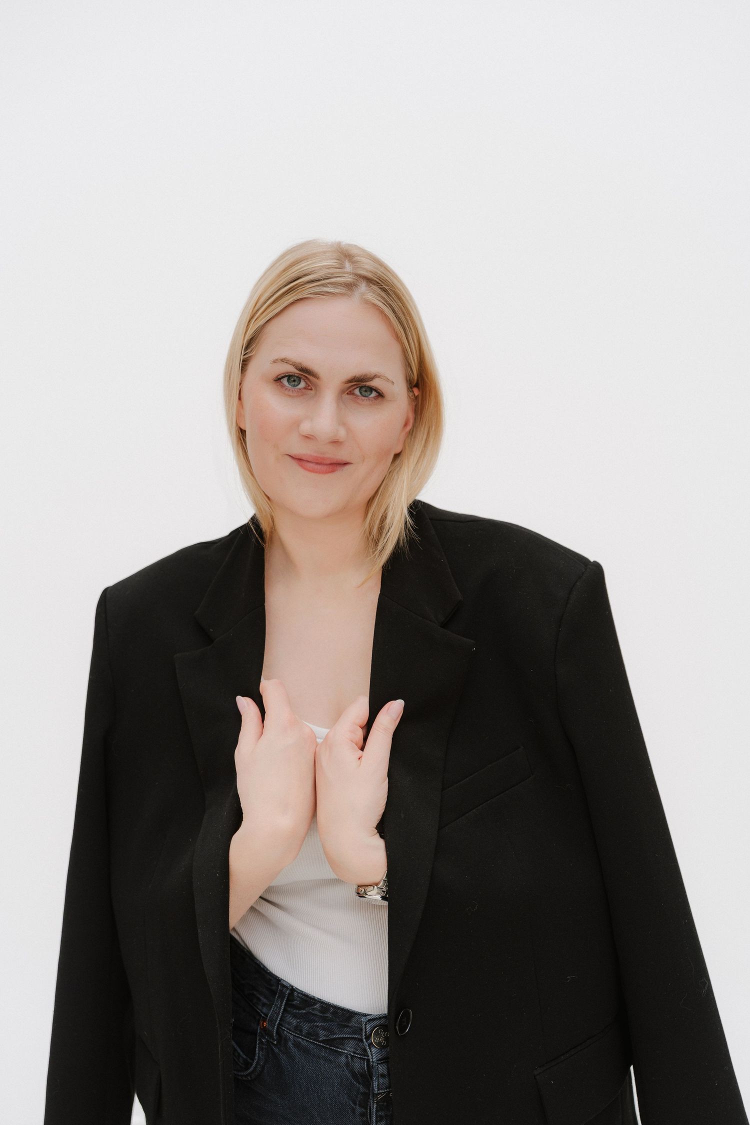 Blonde woman in black blazer and jeans poses with hands together against white background.
