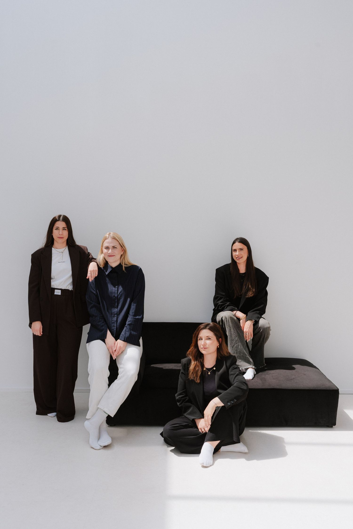 Four women in elegant dark outfits pose confidently against a white wall with a black bench in a modern studio.