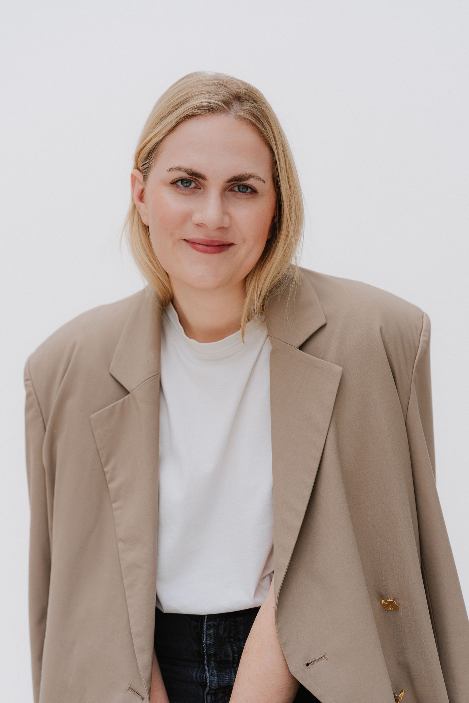 Blonde woman in beige oversized blazer and white t-shirt posing against a light background.