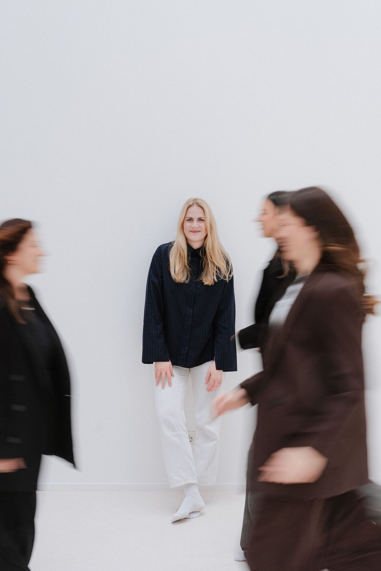 Blonde woman in navy blazer and white pants stands still against white wall while blurred figures walk past her.