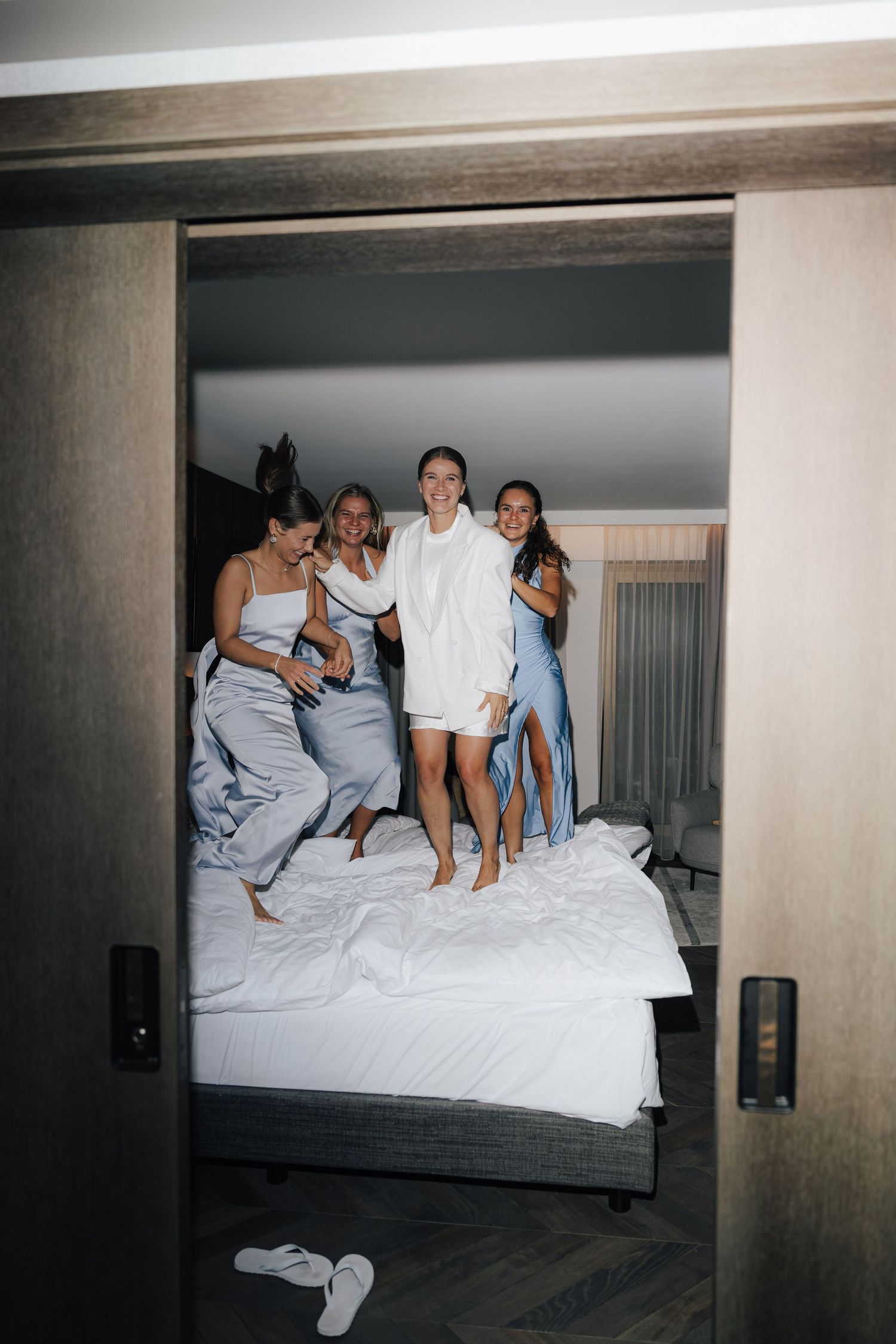 Wedding party jumping and playing on hotel bed in celebratory moment.