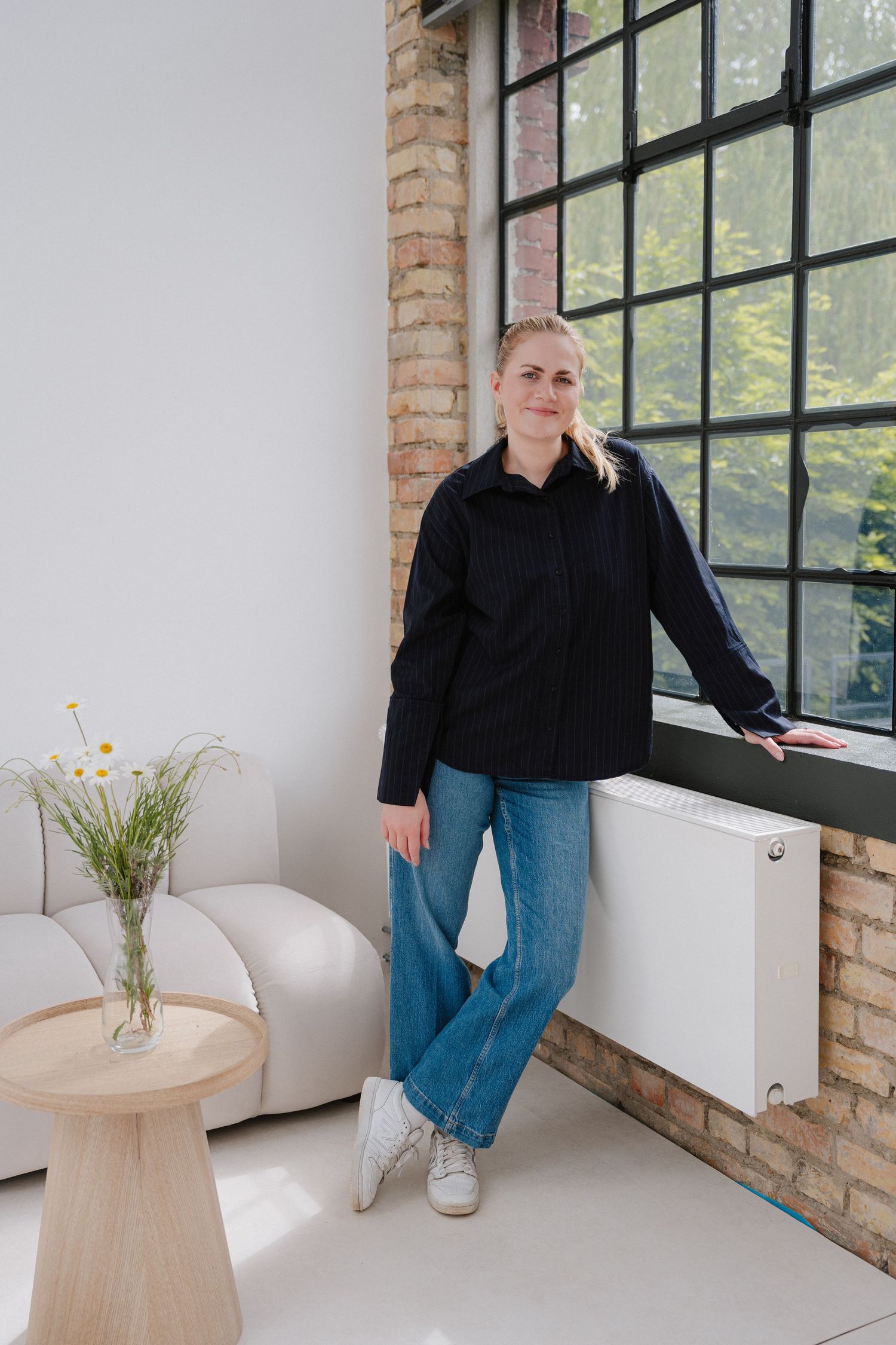 Woman in black shirt and jeans leaning against window in modern loft space with brick wall and white sofa.
