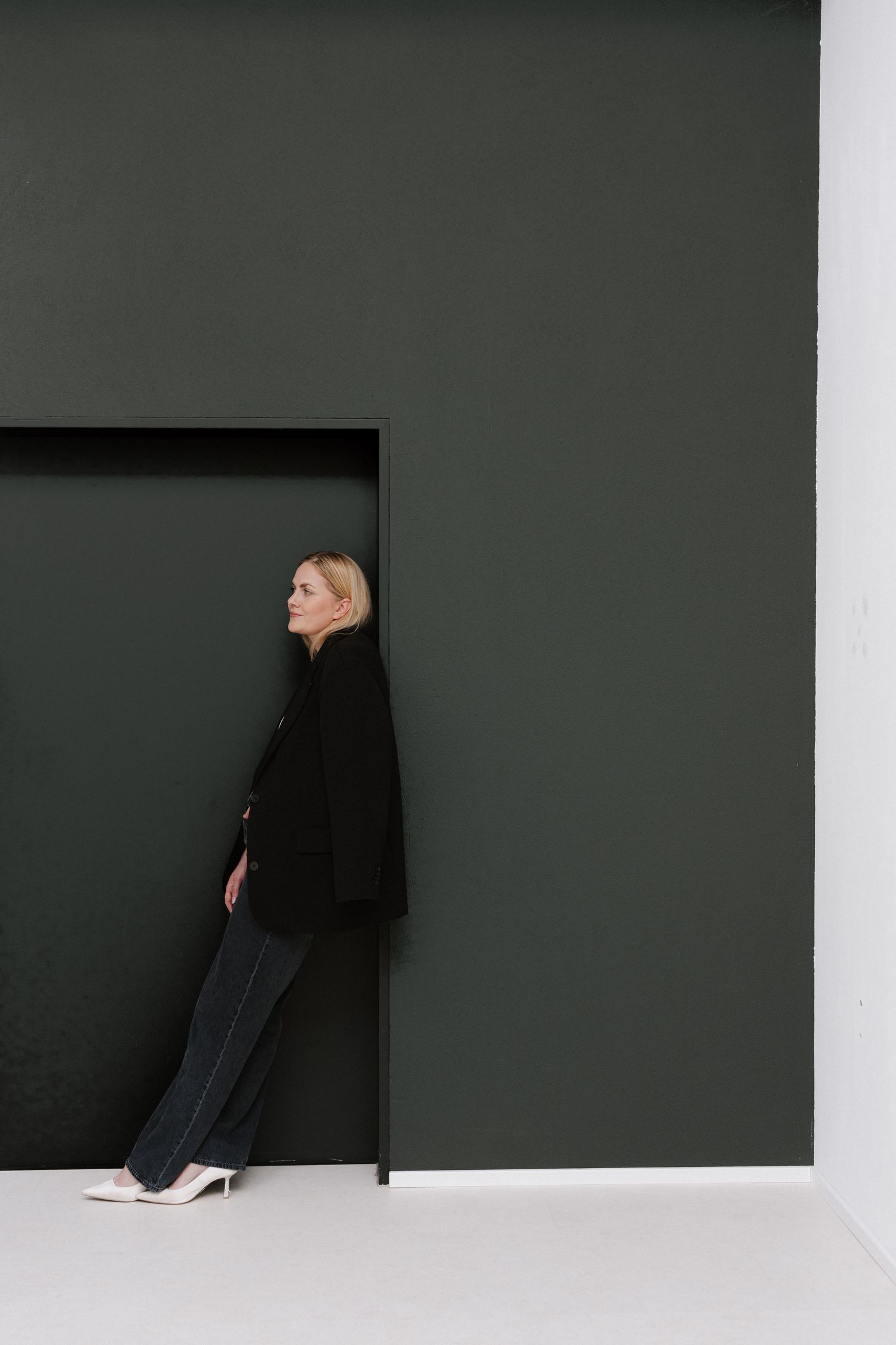A blonde woman in a black blazer and trousers leans against a dark green wall in a minimalist setting.