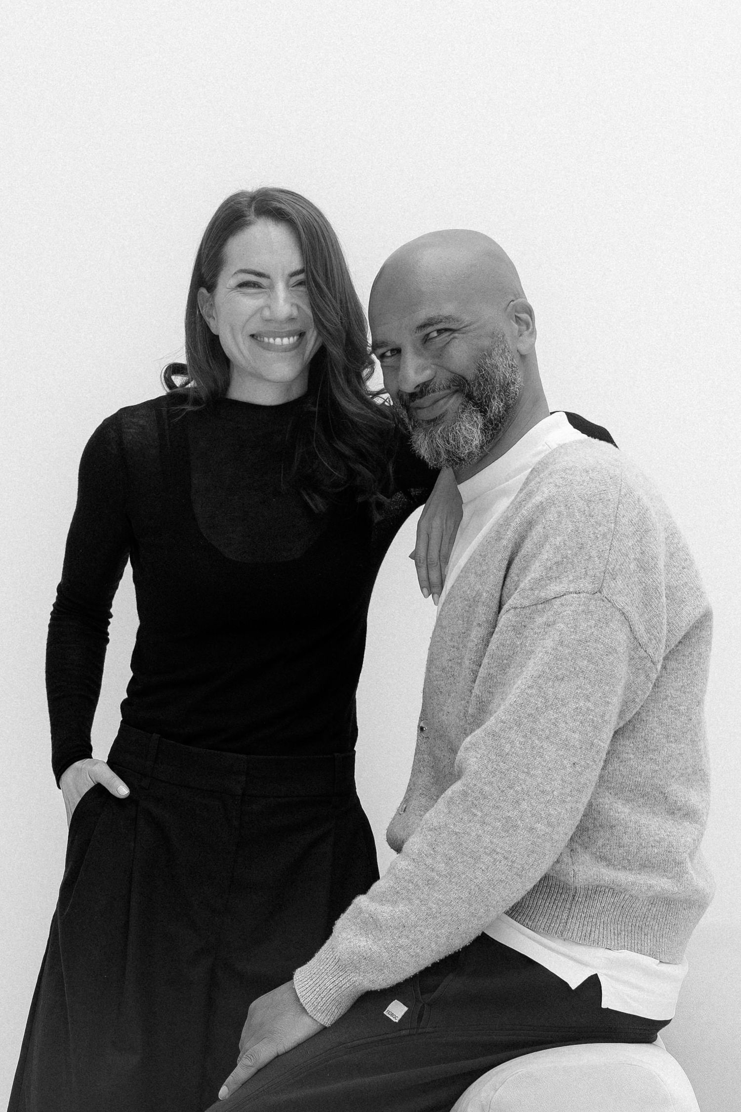 Black and white portrait of a smiling woman in black and a bald bearded man in a light sweater posing together.