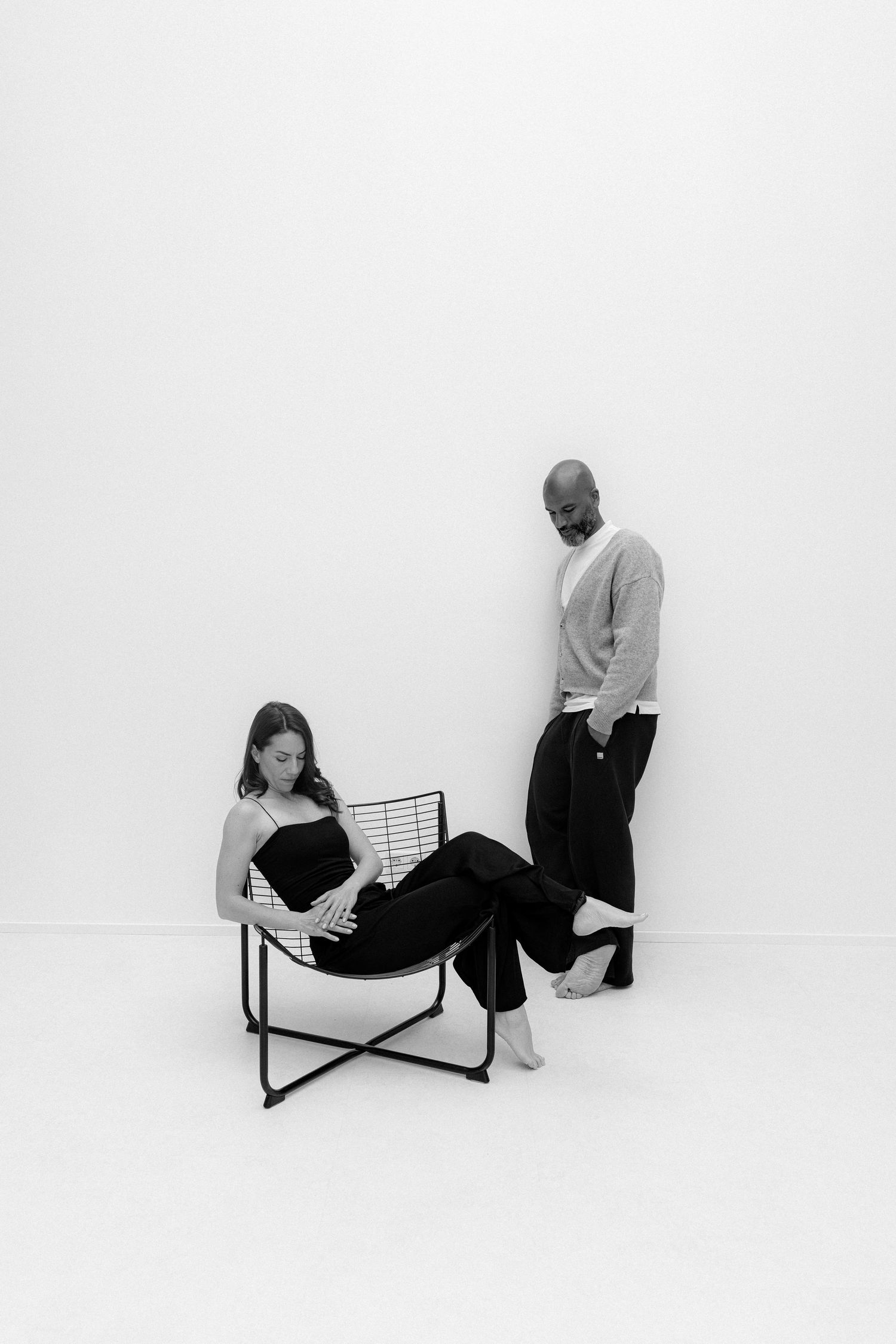 Black and white photo of a woman sitting in a wire chair and a man standing beside her in a minimalist studio.