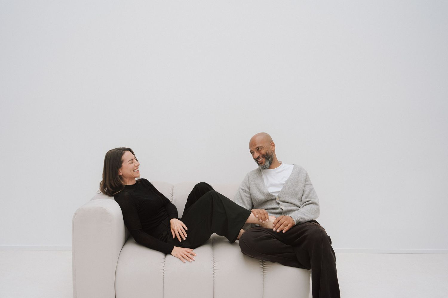 Two people relaxing on a white sofa, smiling and conversing in a minimalist bright room.