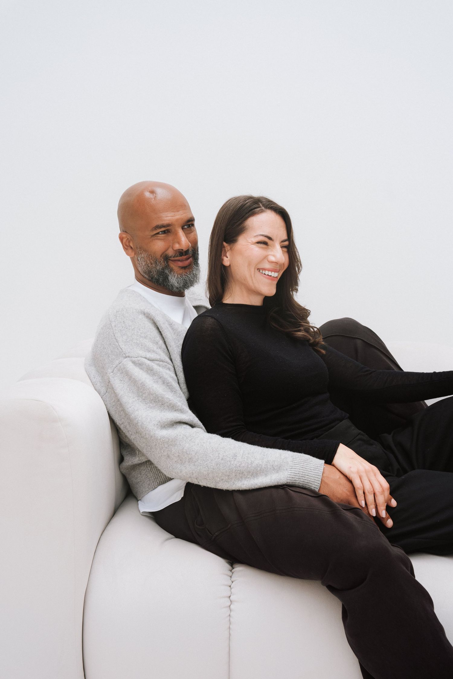 A smiling couple sits together on a white sofa, man in gray sweater and woman in black outfit, against a white background.