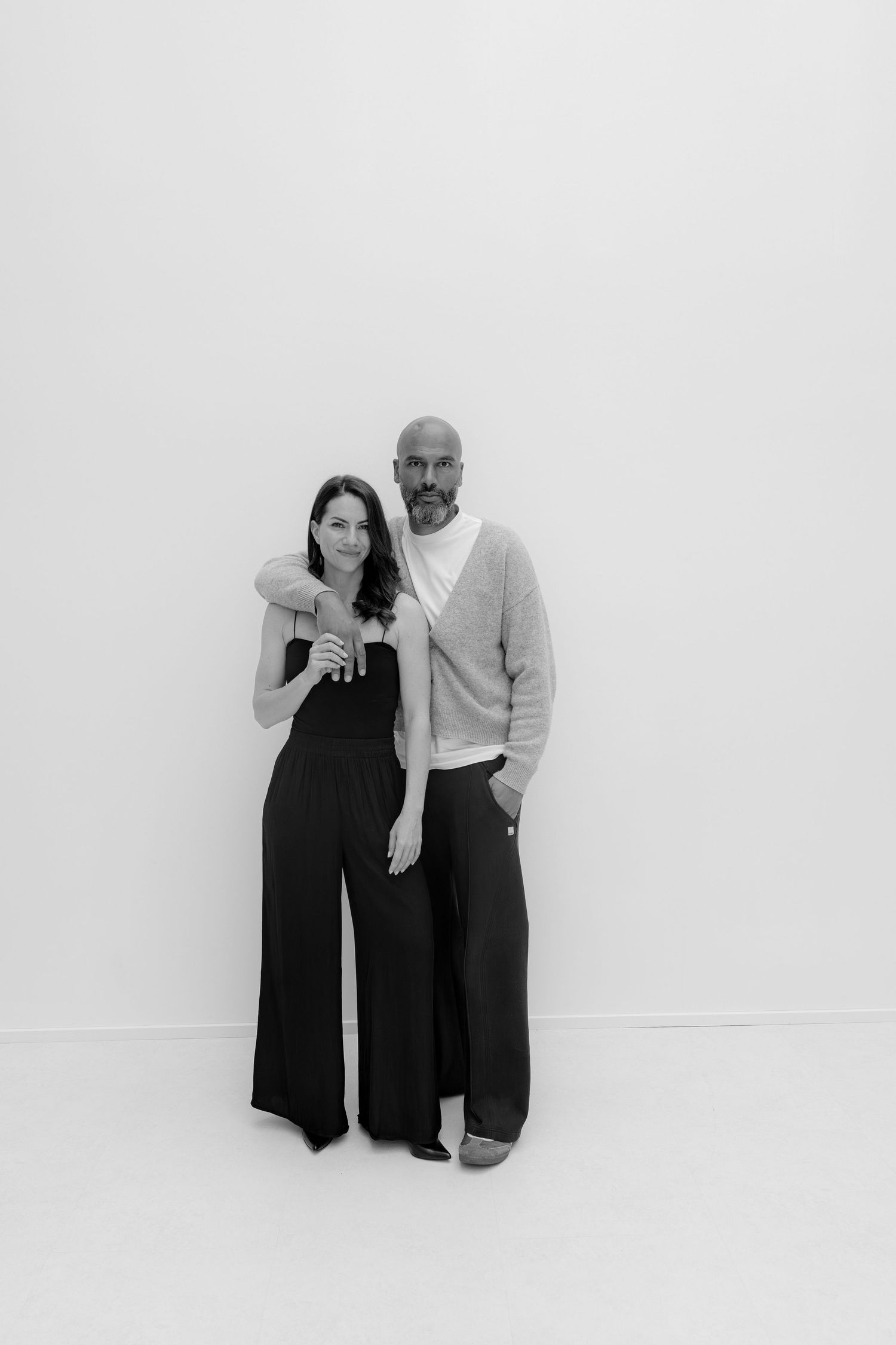 Black and white portrait of a smiling couple standing together against a white background, dressed in elegant dark outfits.