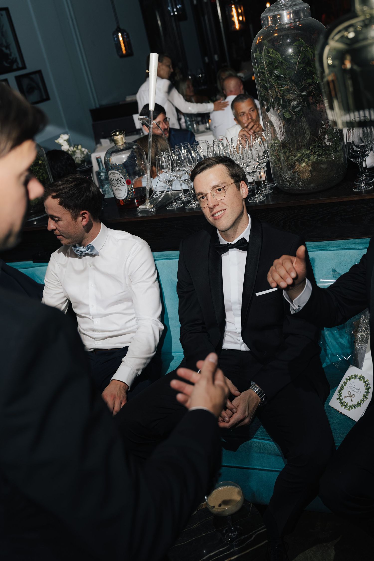 Wedding reception guests in formal attire gathered around tables in a dimly lit venue.