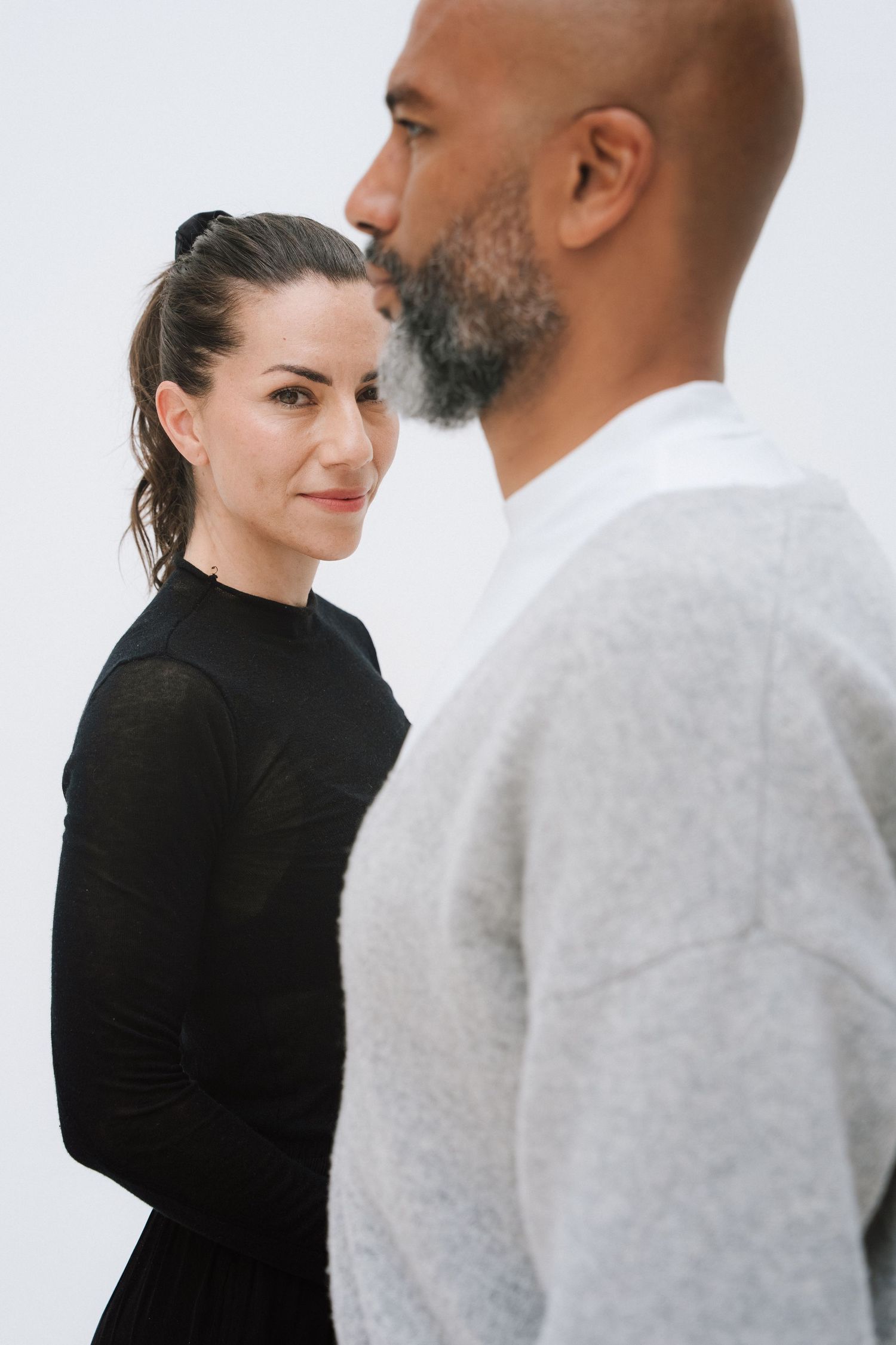 A woman in black smiles at the camera while a bald bearded man in a gray sweater stands in the foreground.