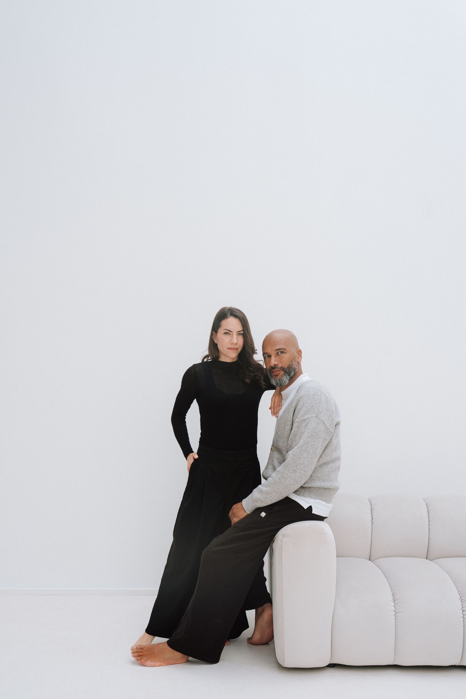 A man and woman pose together in a minimalist white room, she in black and he in gray beside a white sofa.