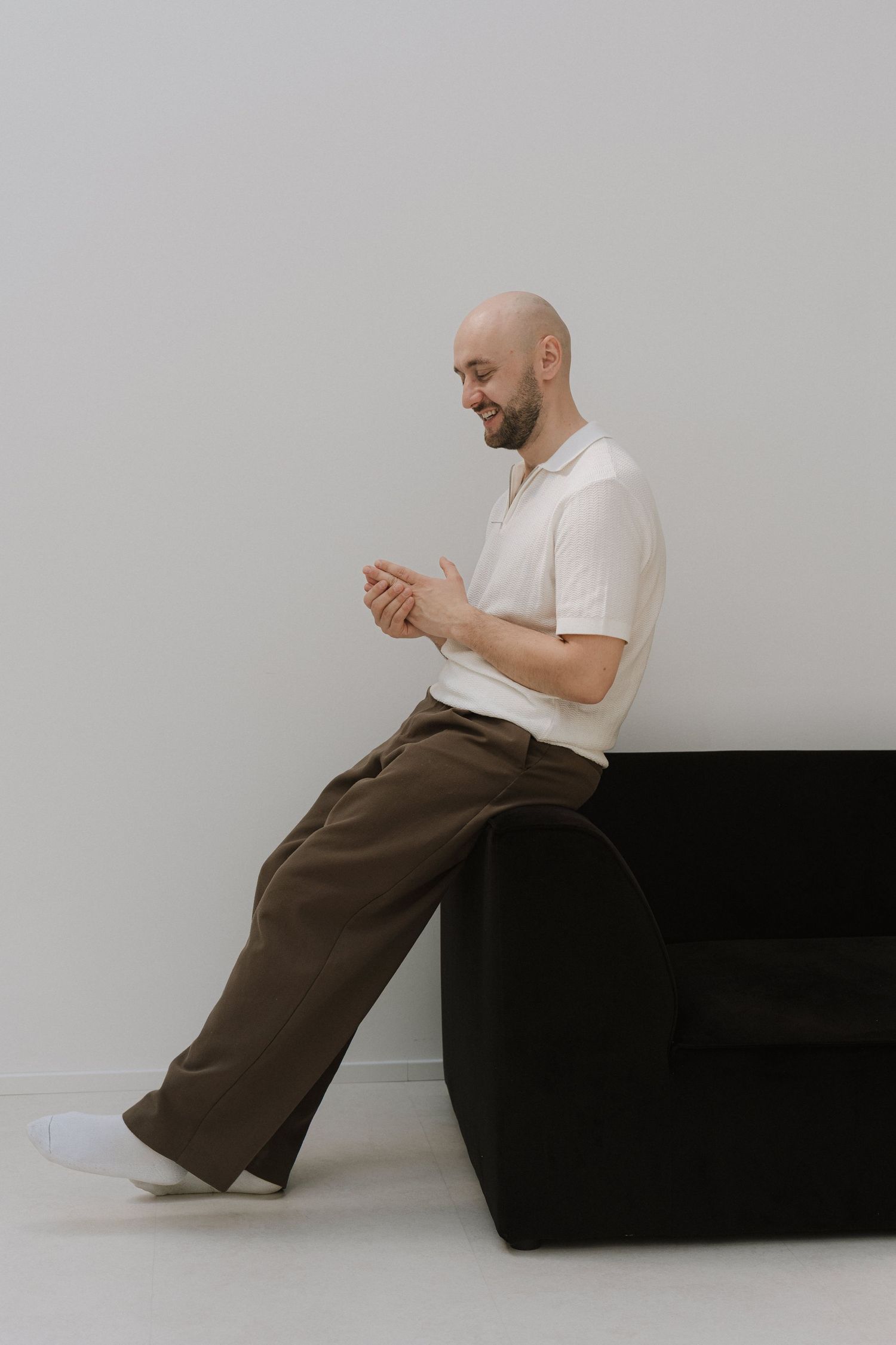 Bald man in white polo and brown pants sitting on black sofa, smiling while looking at his hands.