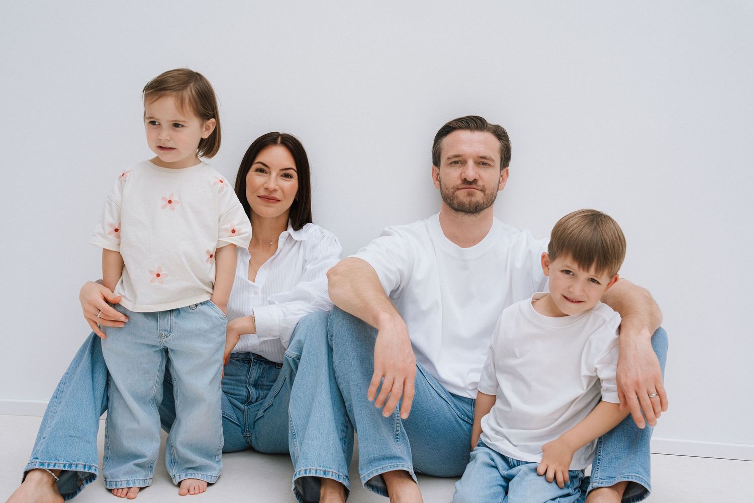 Family of four in white tops and jeans sitting together against a white wall, smiling warmly for a portrait.