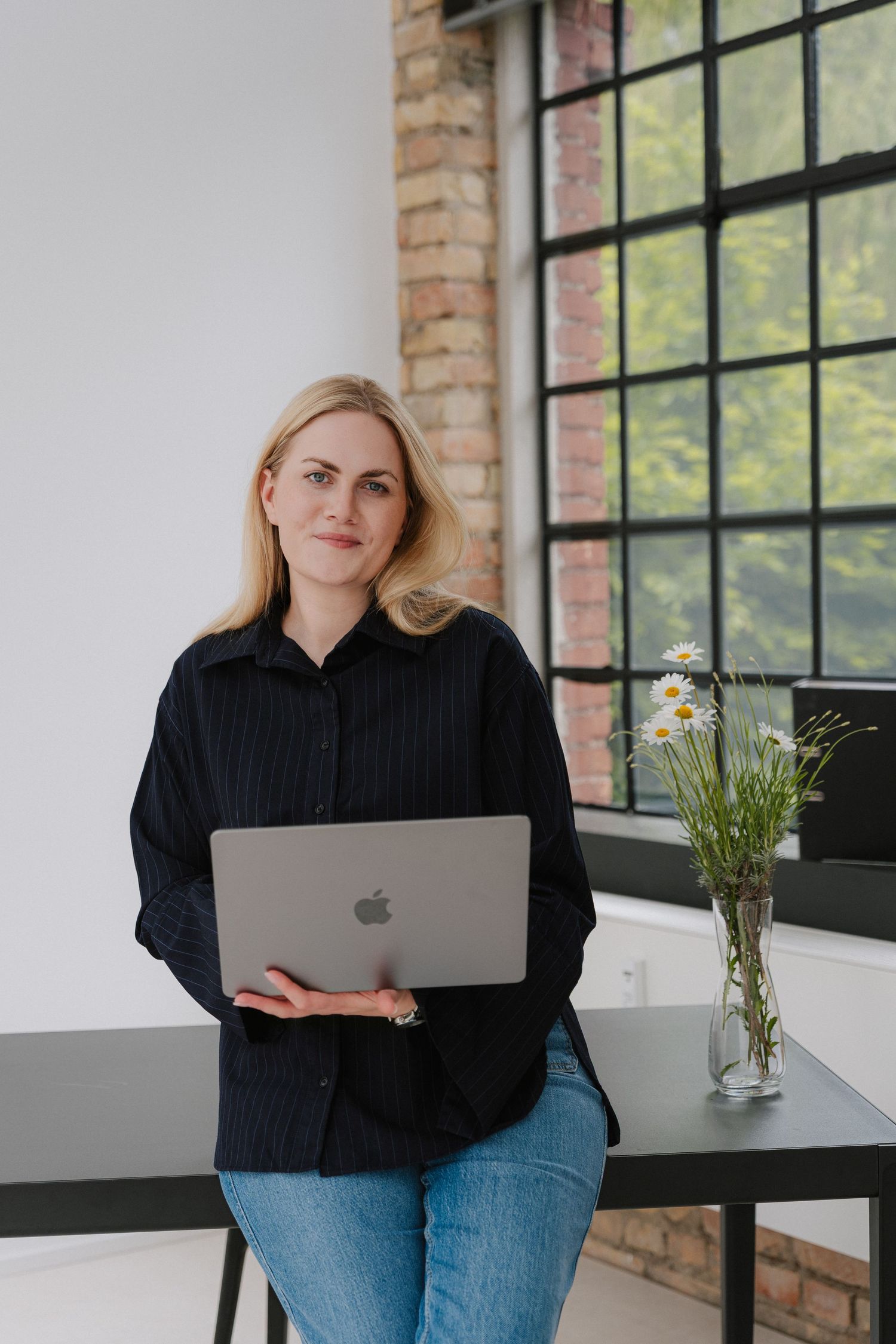 Woman in black shirt holds Apple MacBook laptop, leaning against desk near large industrial windows with daisies.
