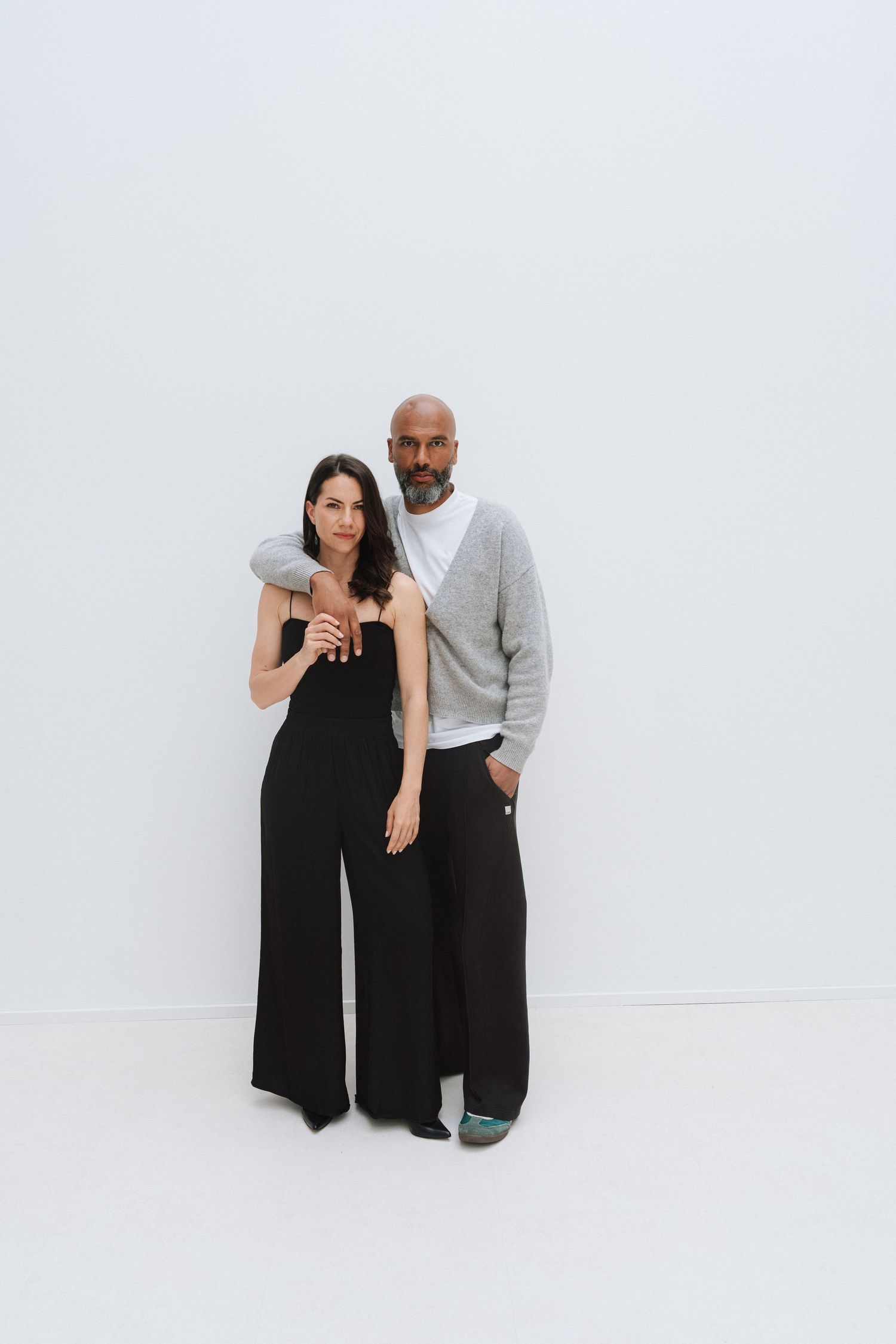 A woman in black wide-leg jumpsuit and a bald man in gray sweater pose together against a white background.