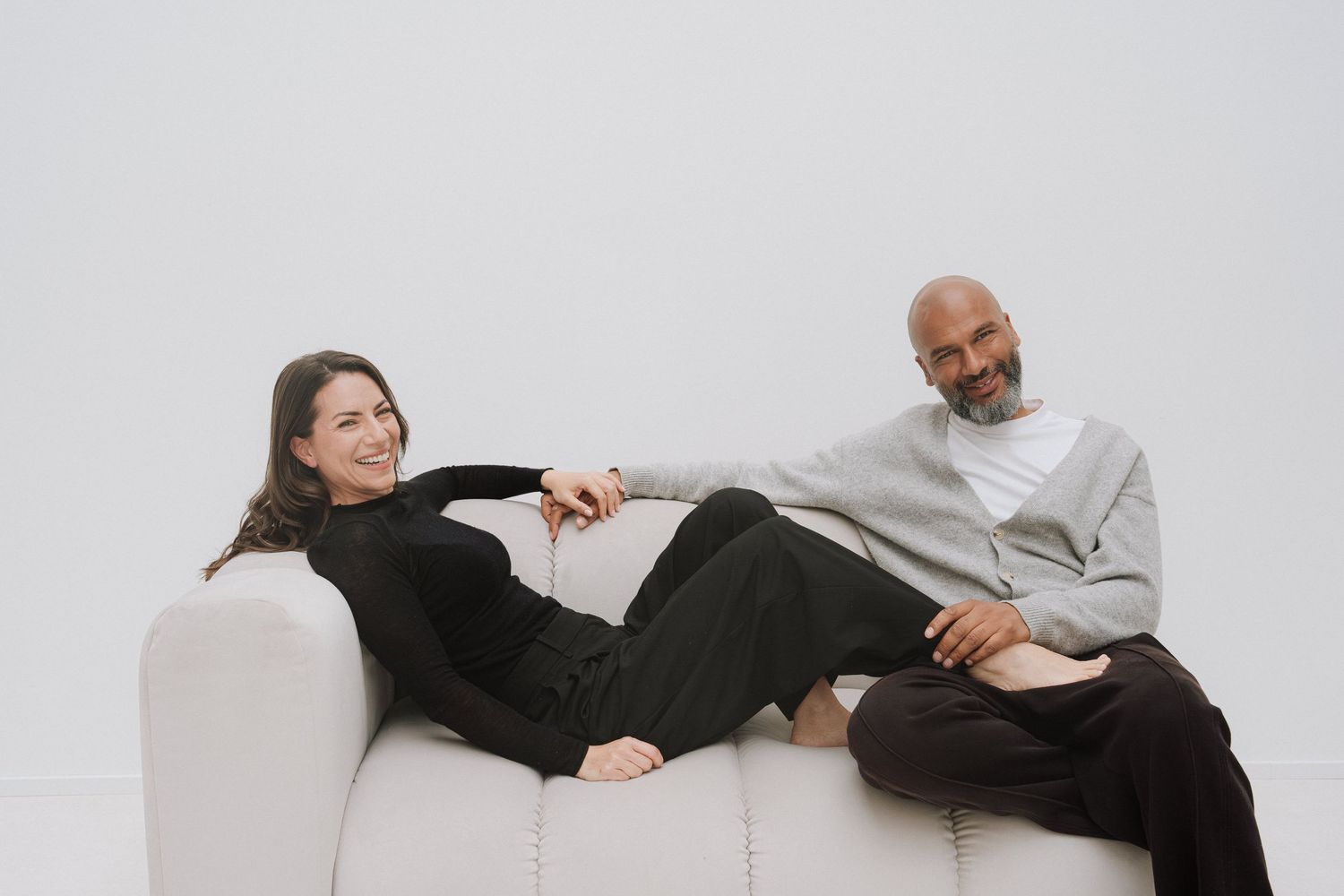 A woman and man sitting together on a white sofa, smiling in a bright minimalist studio setting.