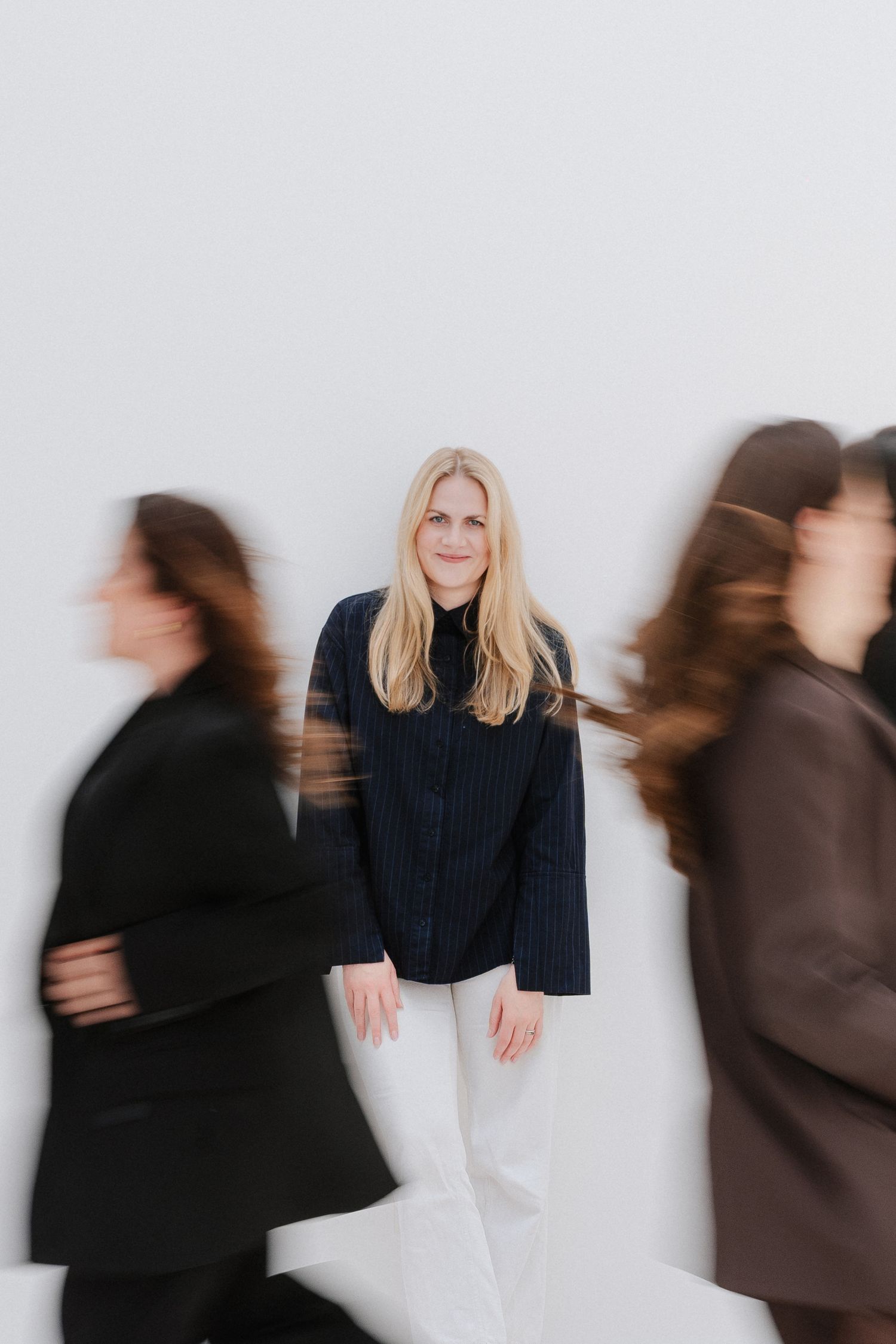 Blonde woman stands still in dark blue shirt and white pants while blurred figures rush past on both sides.