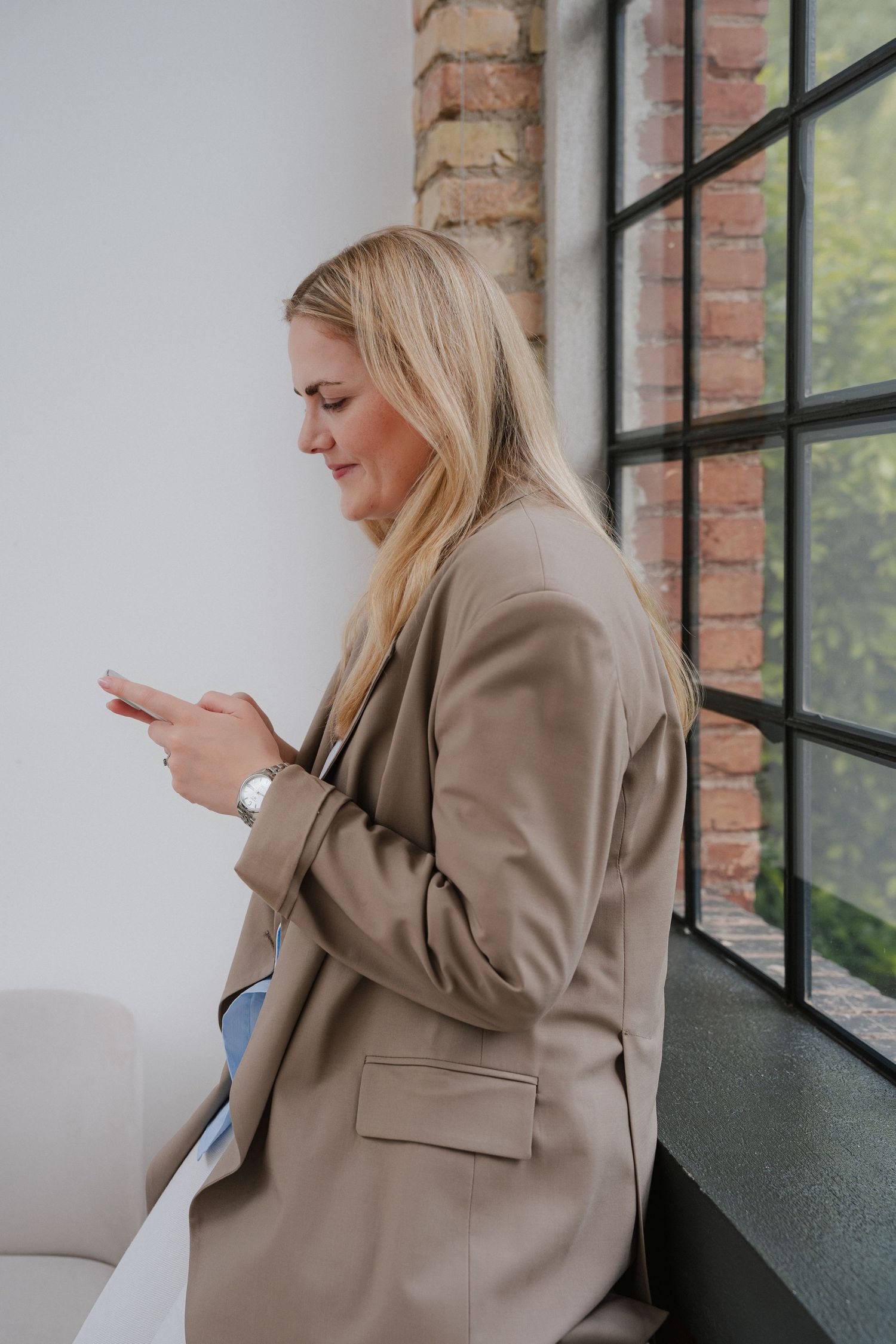 Blonde woman in beige blazer smiling at her smartphone while sitting by a large window with brick wall backdrop.