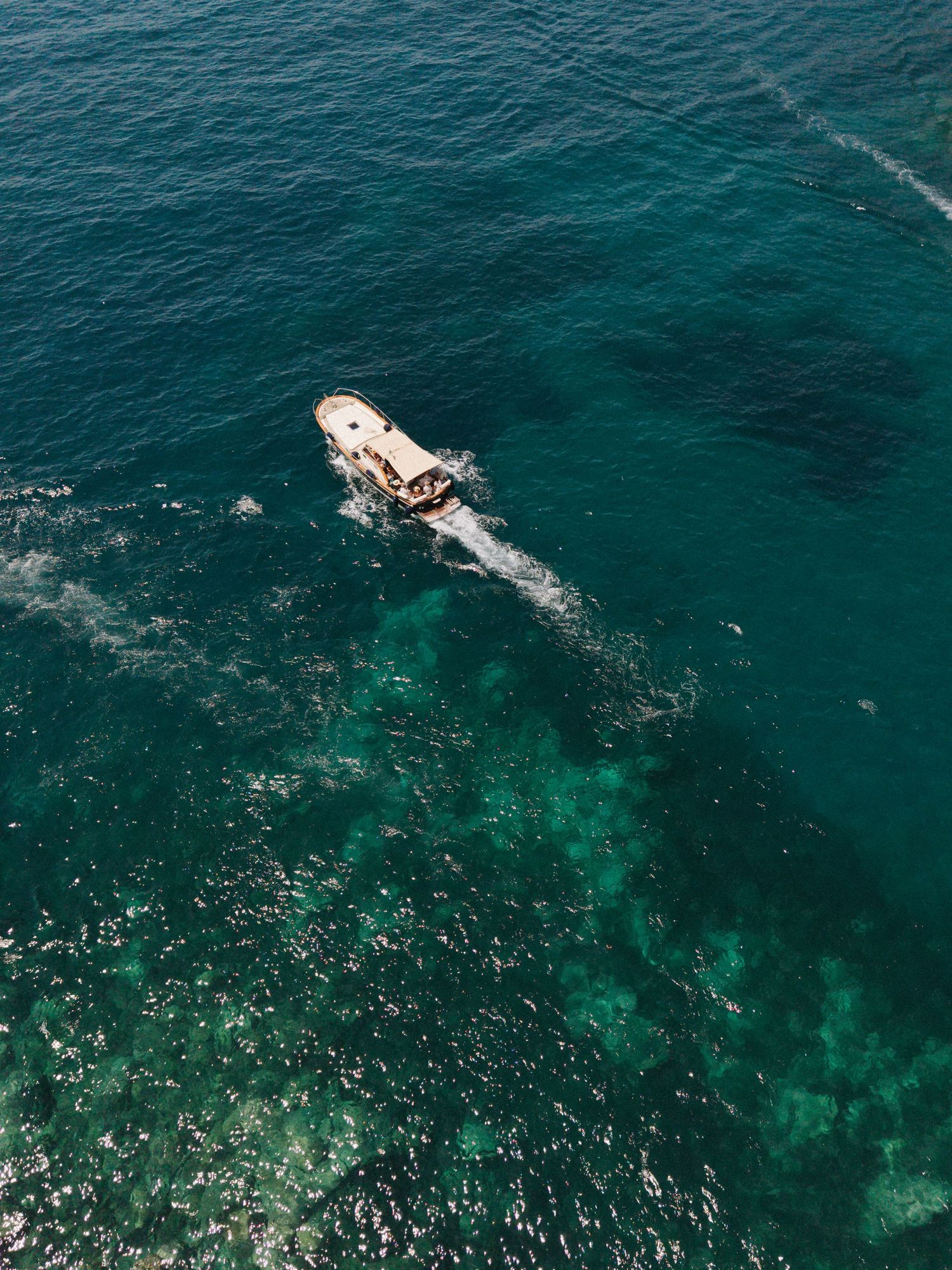 Aerial view of a small boat sailing through clear turquoise ocean water above a coral reef.