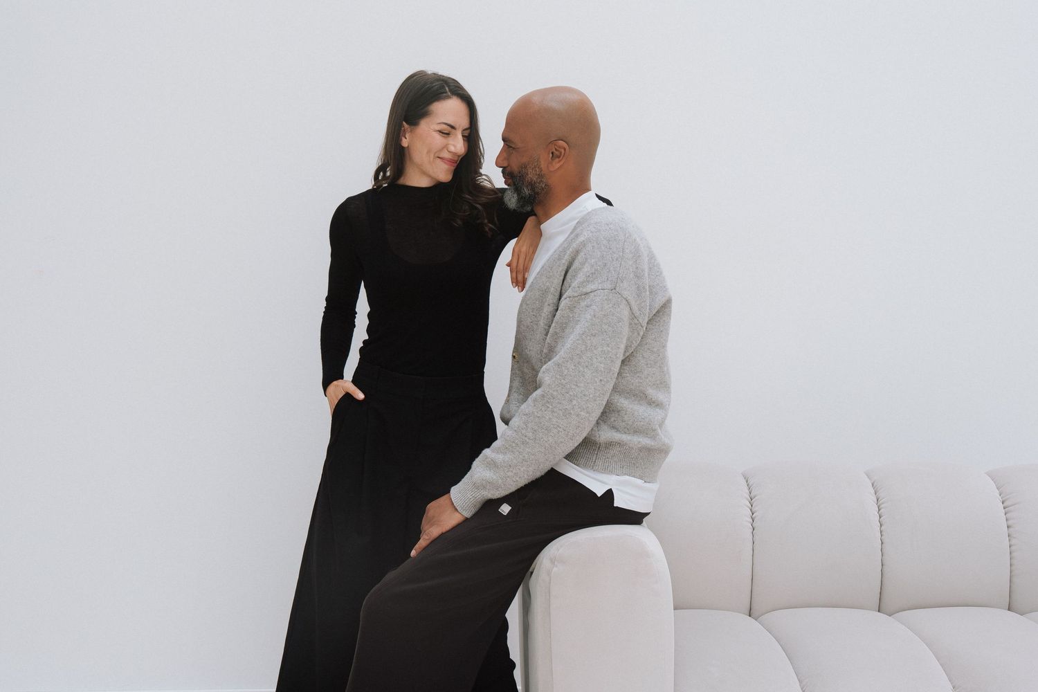 A woman in a black dress and a man in a gray cardigan sit together on a white sofa against a white wall.
