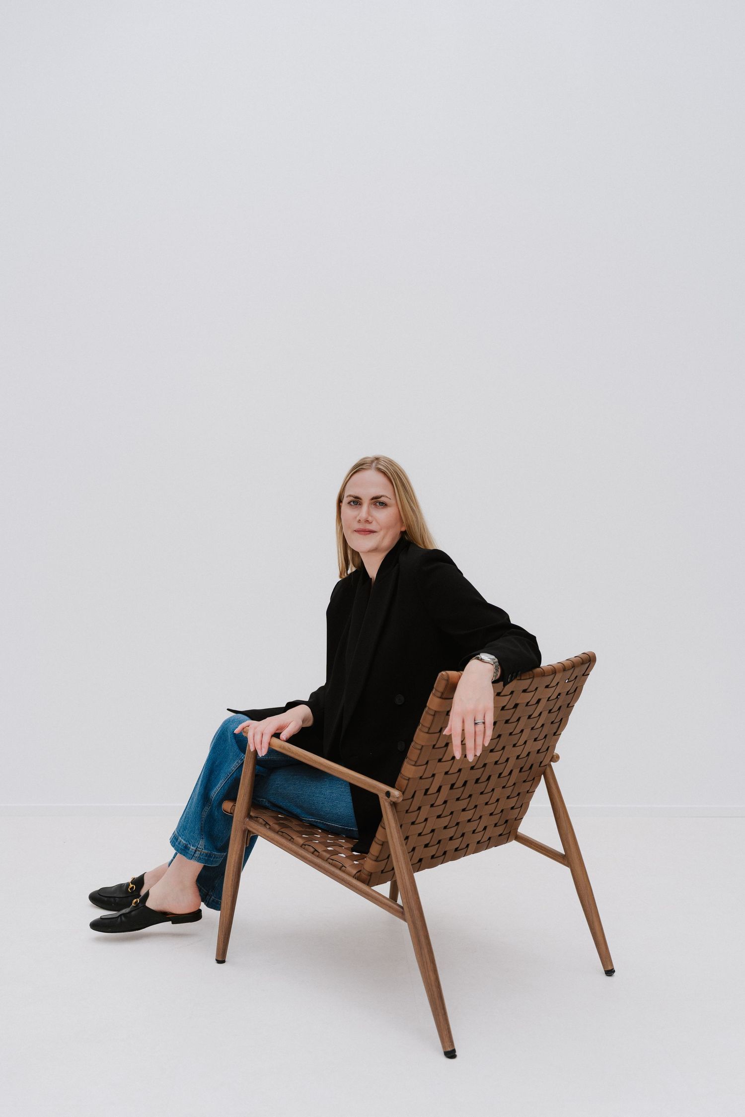 Woman in black blazer and jeans sitting relaxed on a woven wooden chair against a white background.