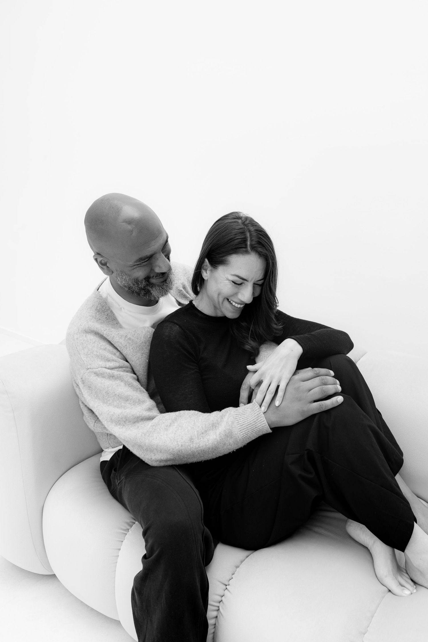Black and white photo of a happy couple embracing on a white sofa, woman smiling with knees drawn up.