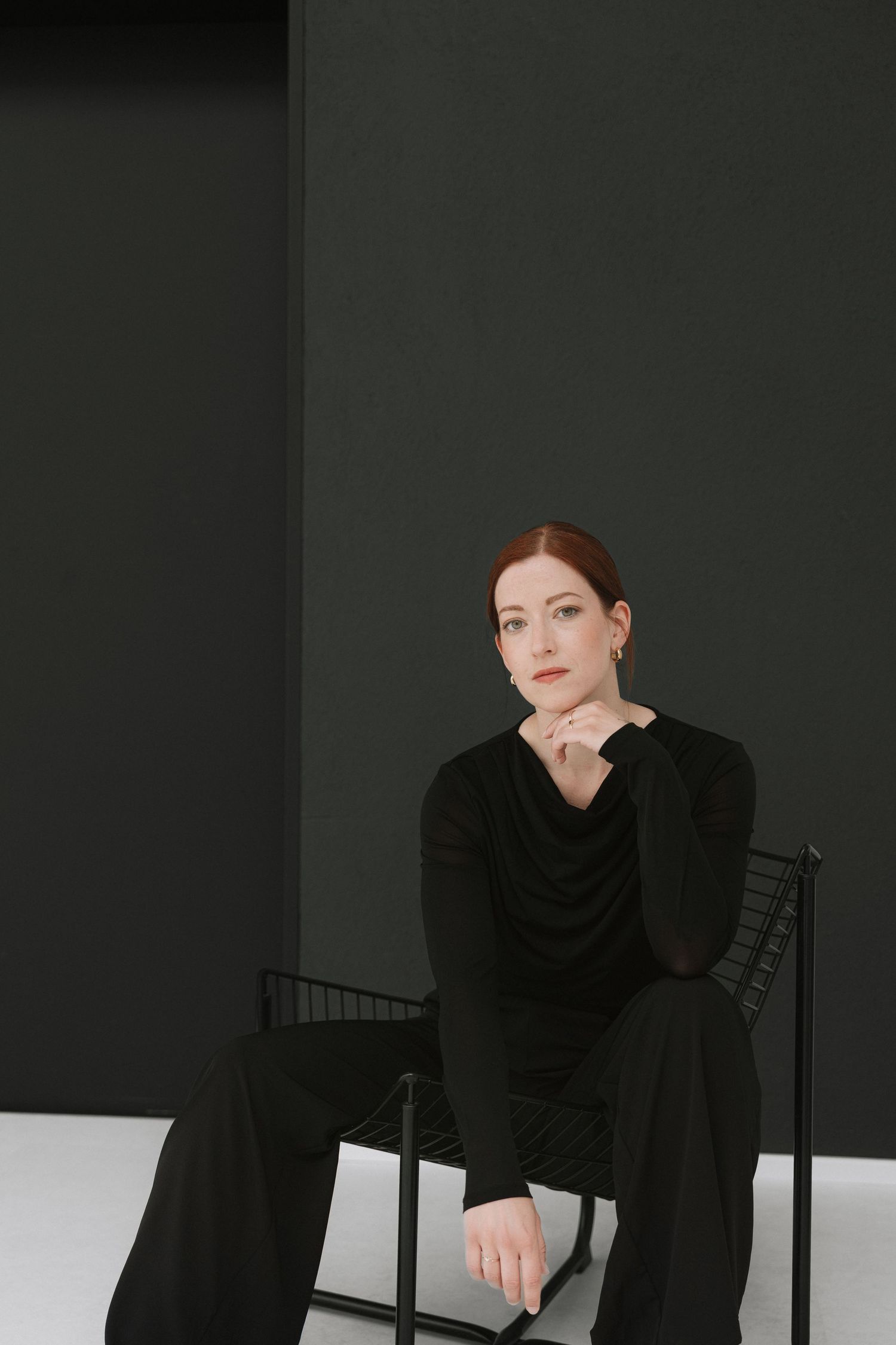 Woman in black outfit sitting on chair against dark green wall, posing with hand on chin.