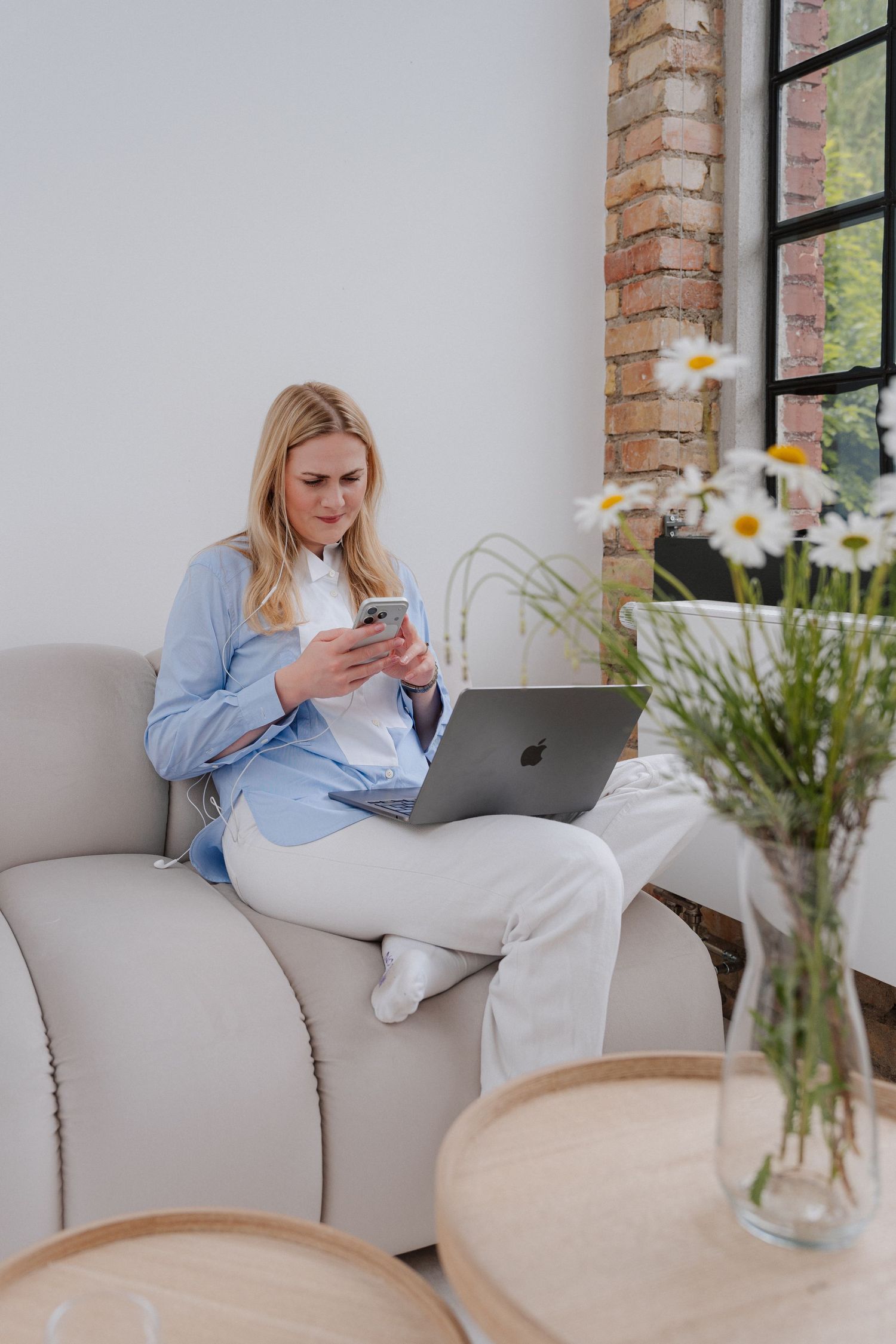 Woman in blue blazer sitting on sofa using smartphone with laptop, beside daisy flowers in modern living room.