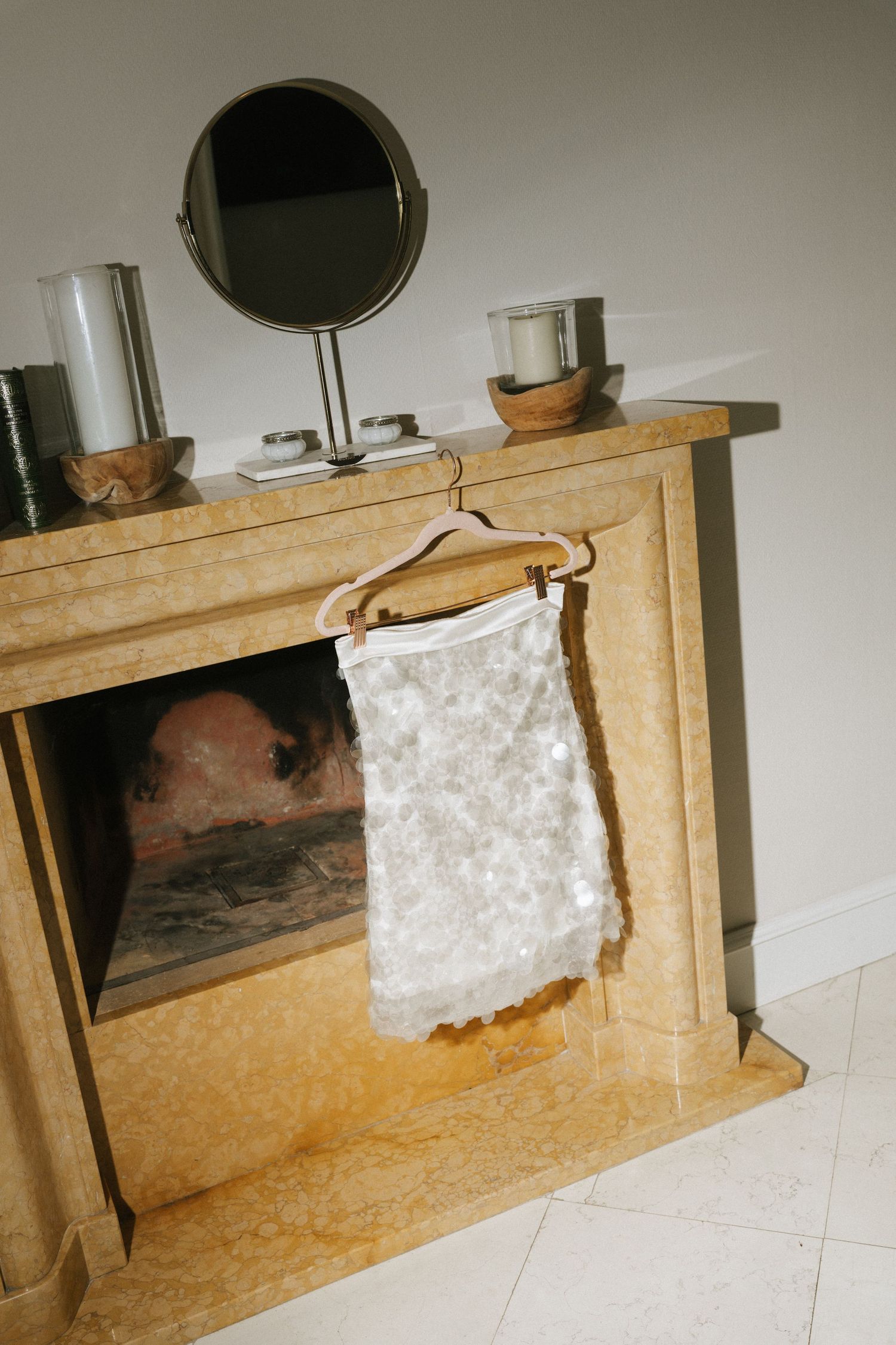 White lace garment on hanger displayed over marble fireplace with round mirror and candles on mantel.