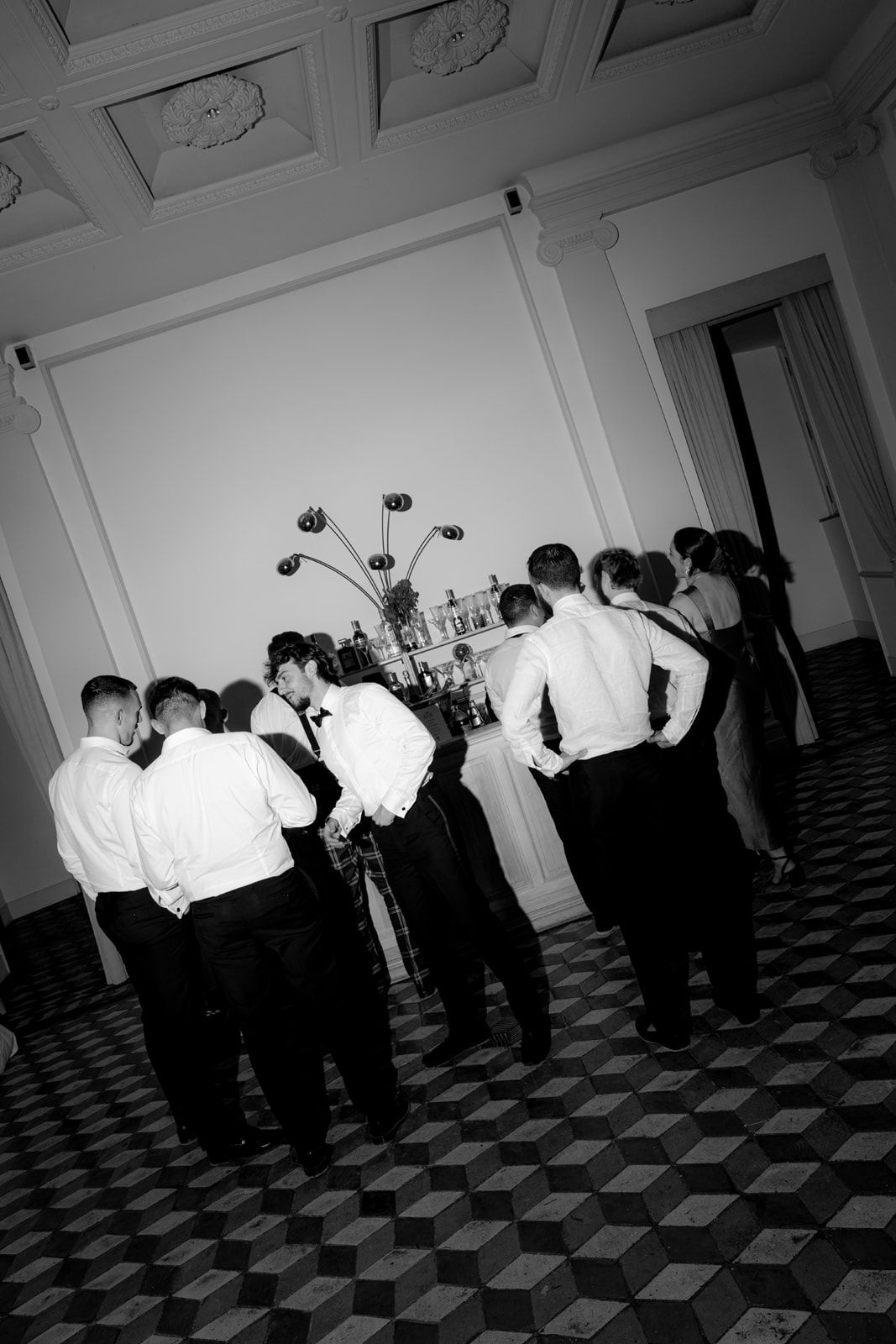Black and white photo of groomsmen in formal attire gathered at a bar in an elegant ballroom.
