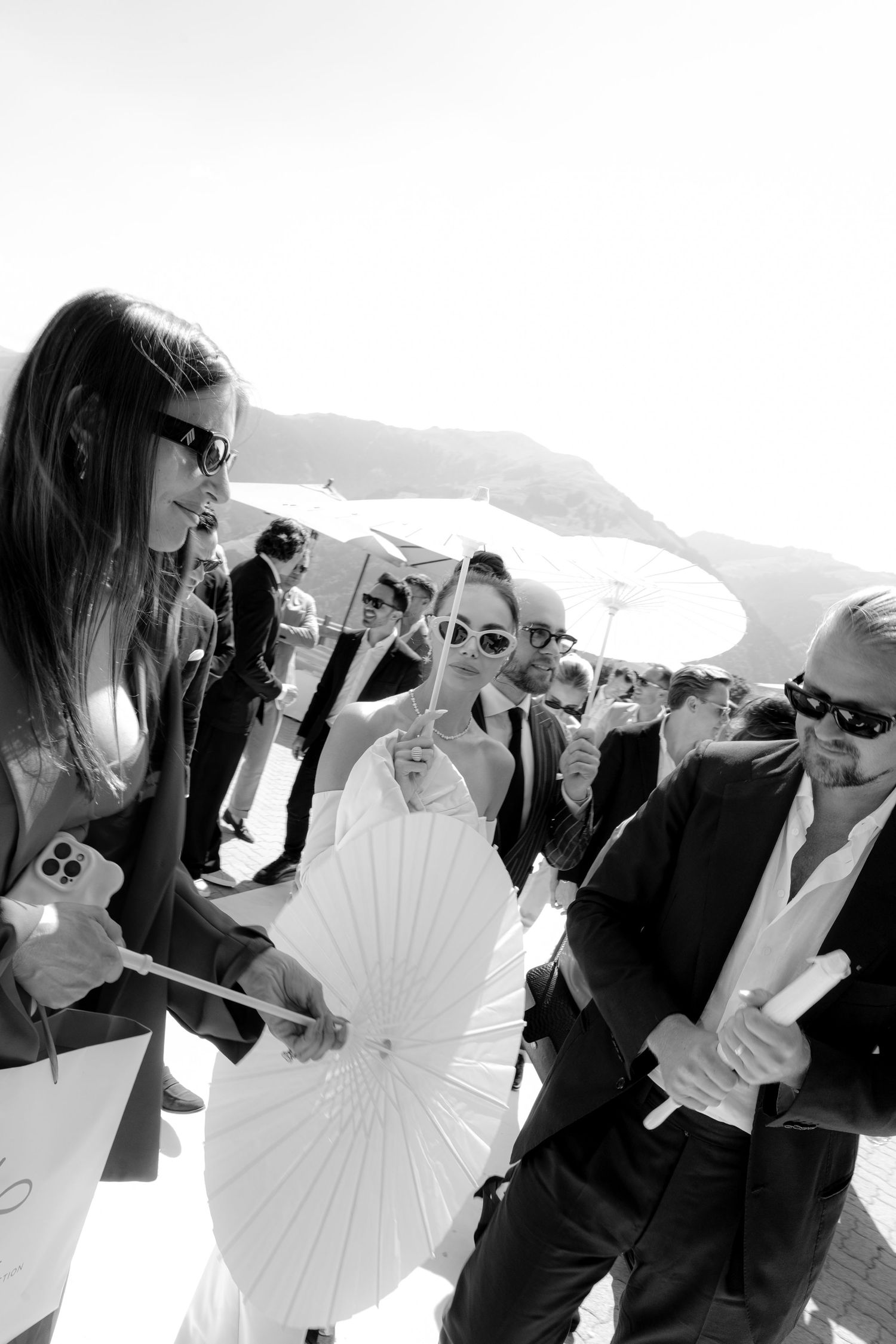 Wedding guests in sunglasses holding white parasols gather outdoors on a sunny day.