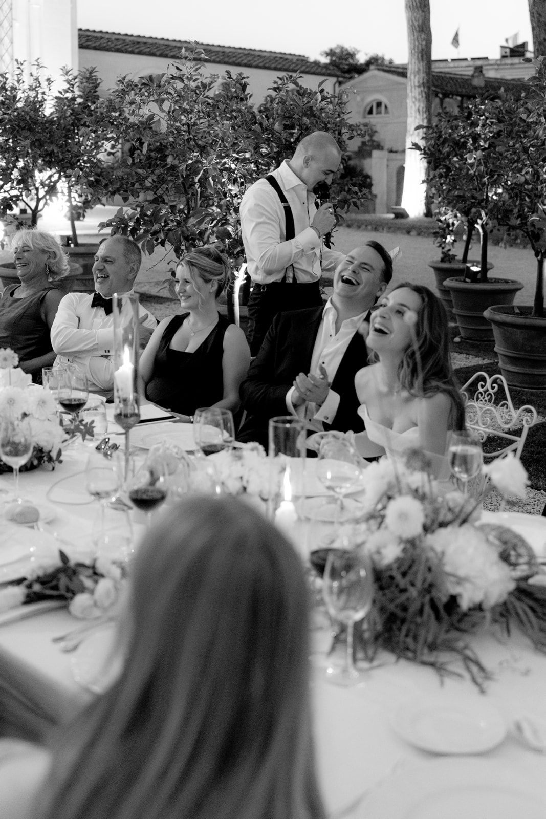 Black and white wedding reception photo of guests laughing at a dinner table with floral centerpieces and candles.