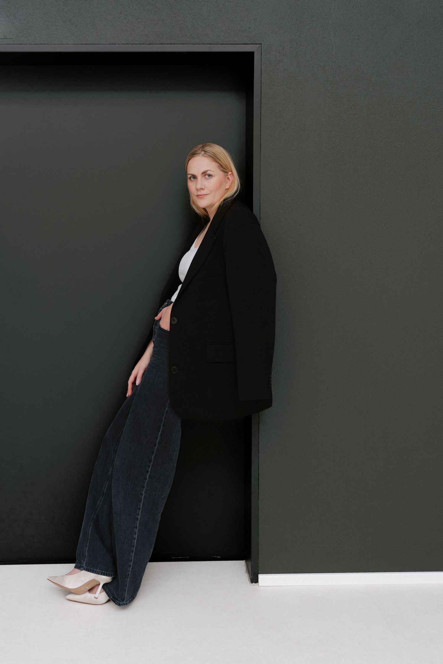 Blonde woman in black blazer and wide-leg jeans leaning against dark green wall in white heels.